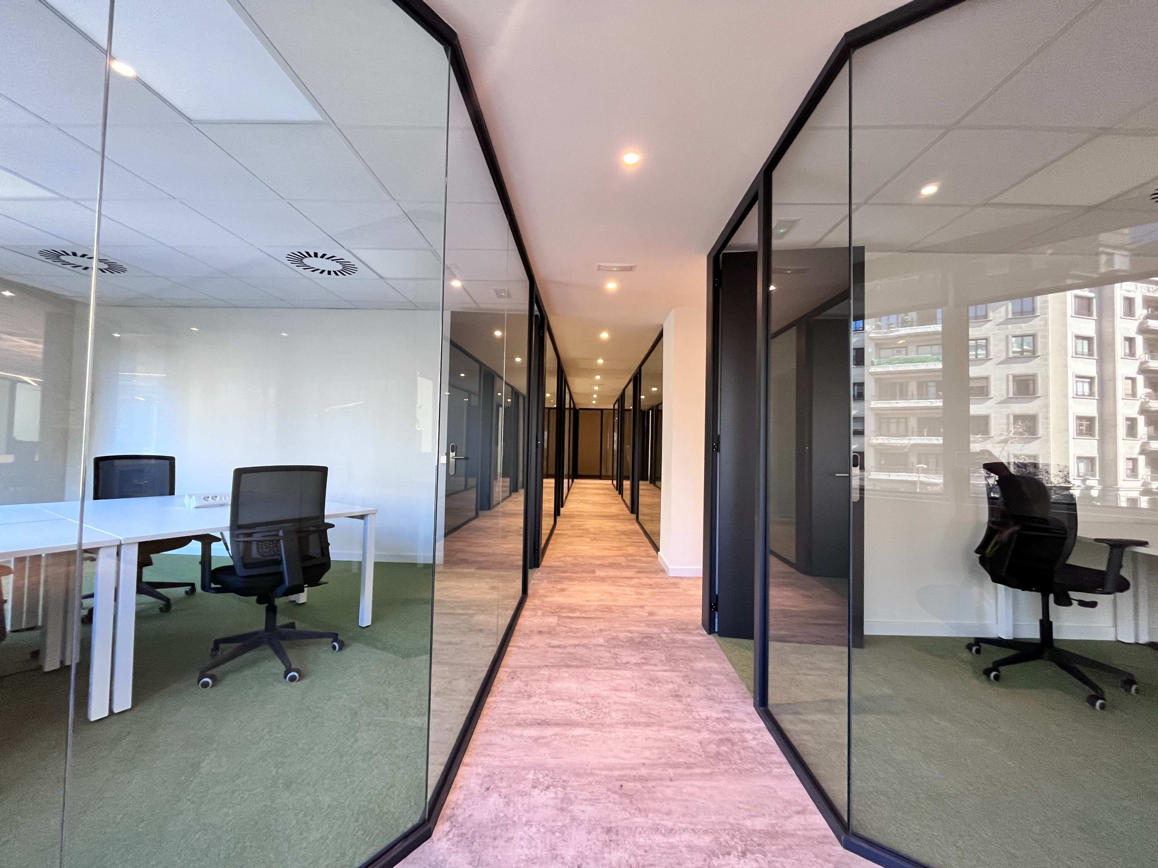 The image shows a modern office corridor lined with glass-walled conference rooms and desks on either side.