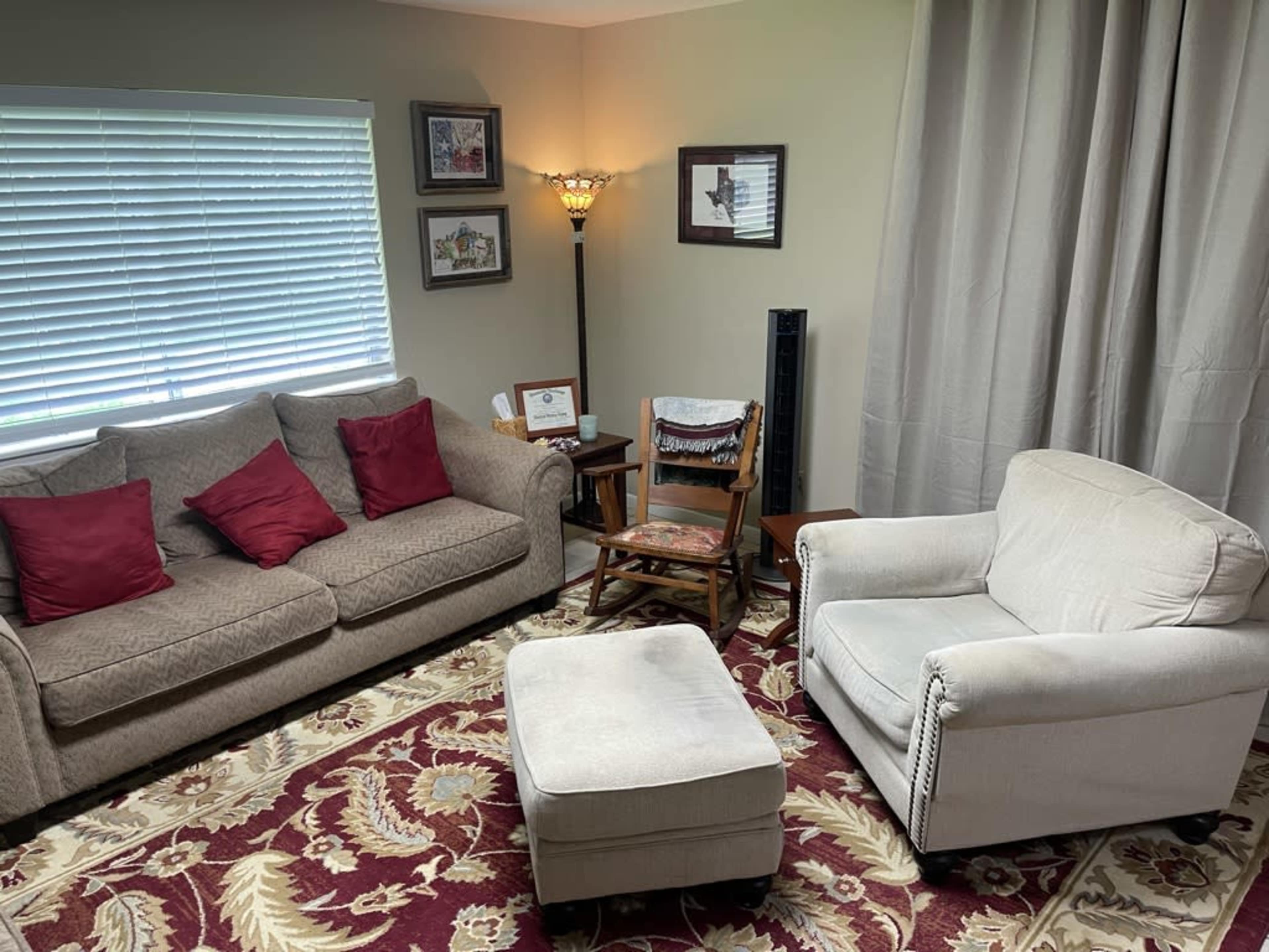 A cozy seating area with a gray sofa, an armchair, an ottoman, a wooden chair, and framed pictures on the wall, all arranged on a patterned rug.