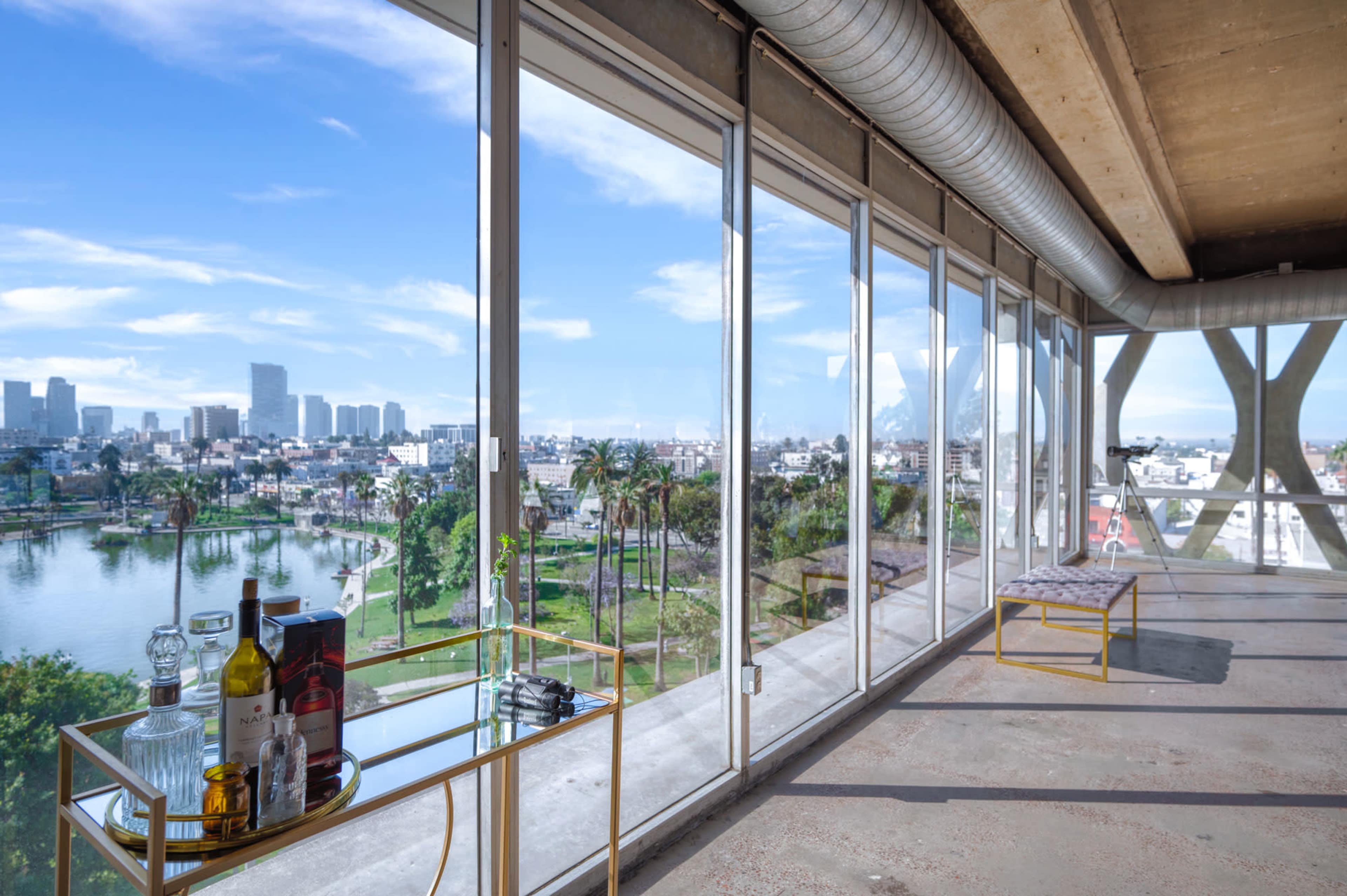A spacious interior with large windows overlooking a cityscape and a body of water, featuring a table with various bottles and glasses.