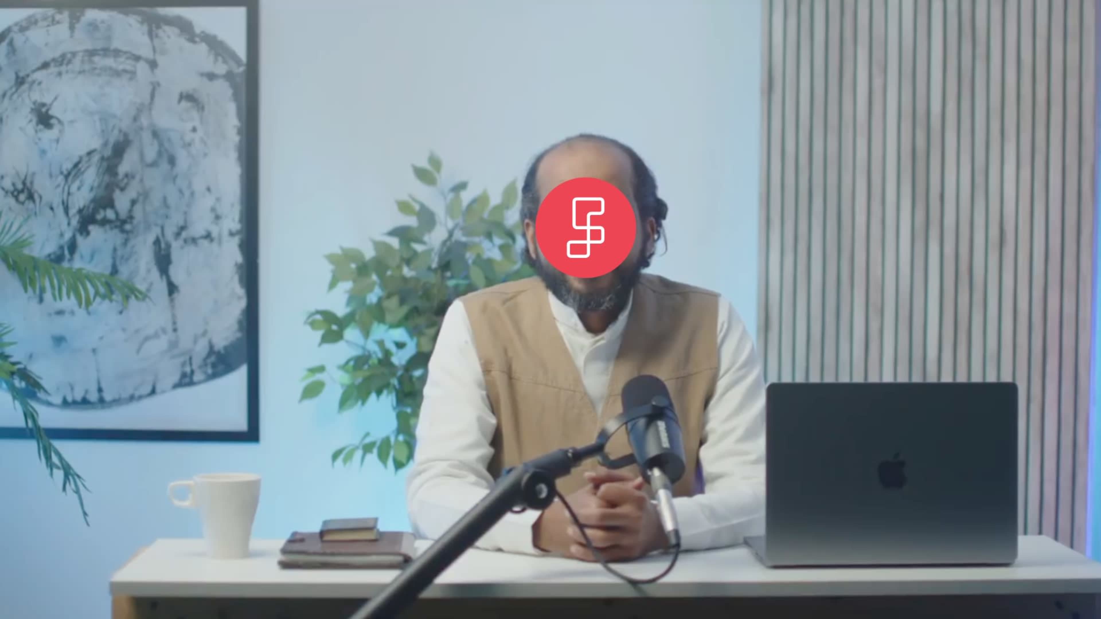 A person sitting at a desk with a microphone and a laptop is surrounded by plants and artwork.