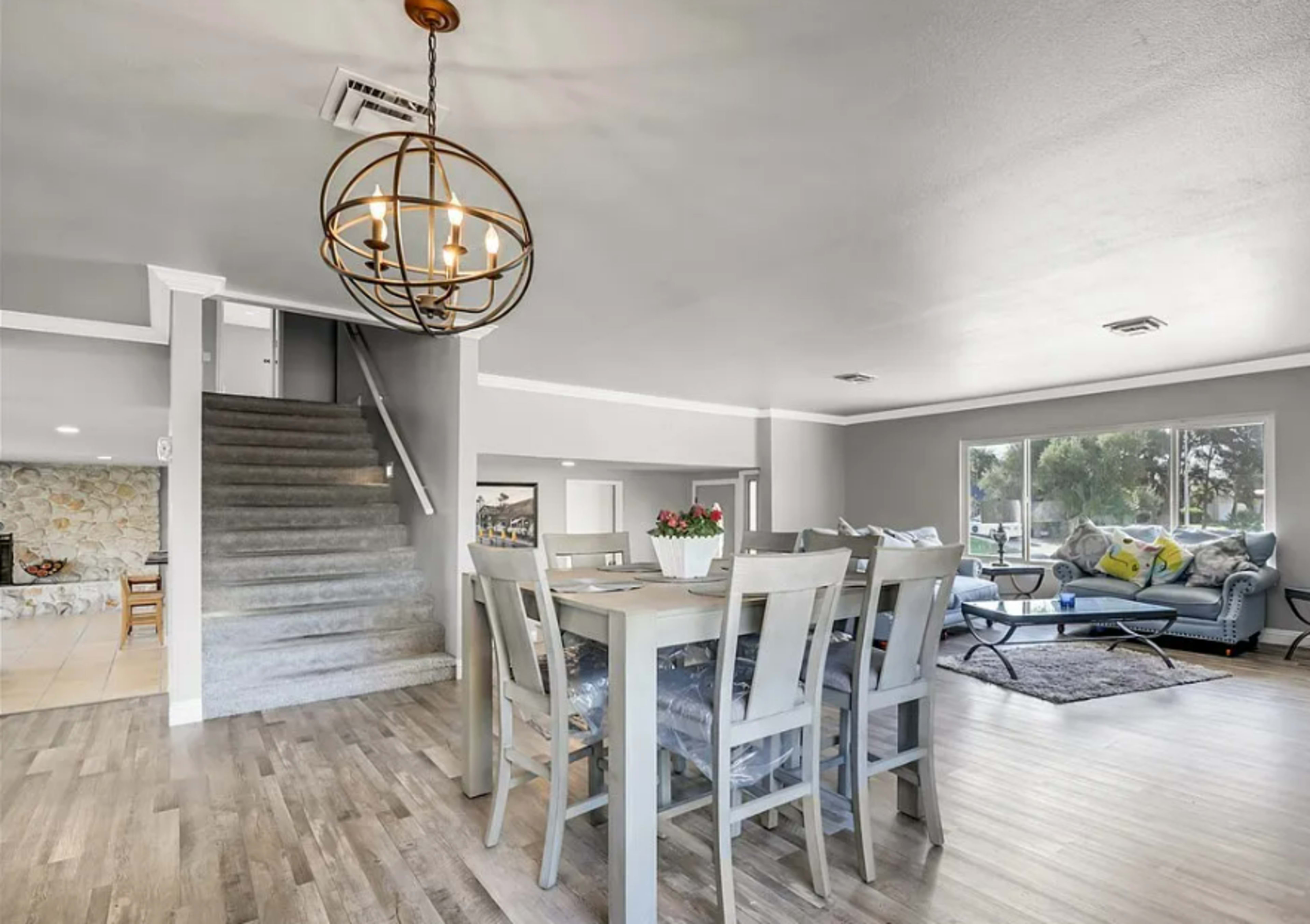 A dining area with a round chandelier and beige wooden table is located adjacent to a living room featuring a blue sofa and large windows.