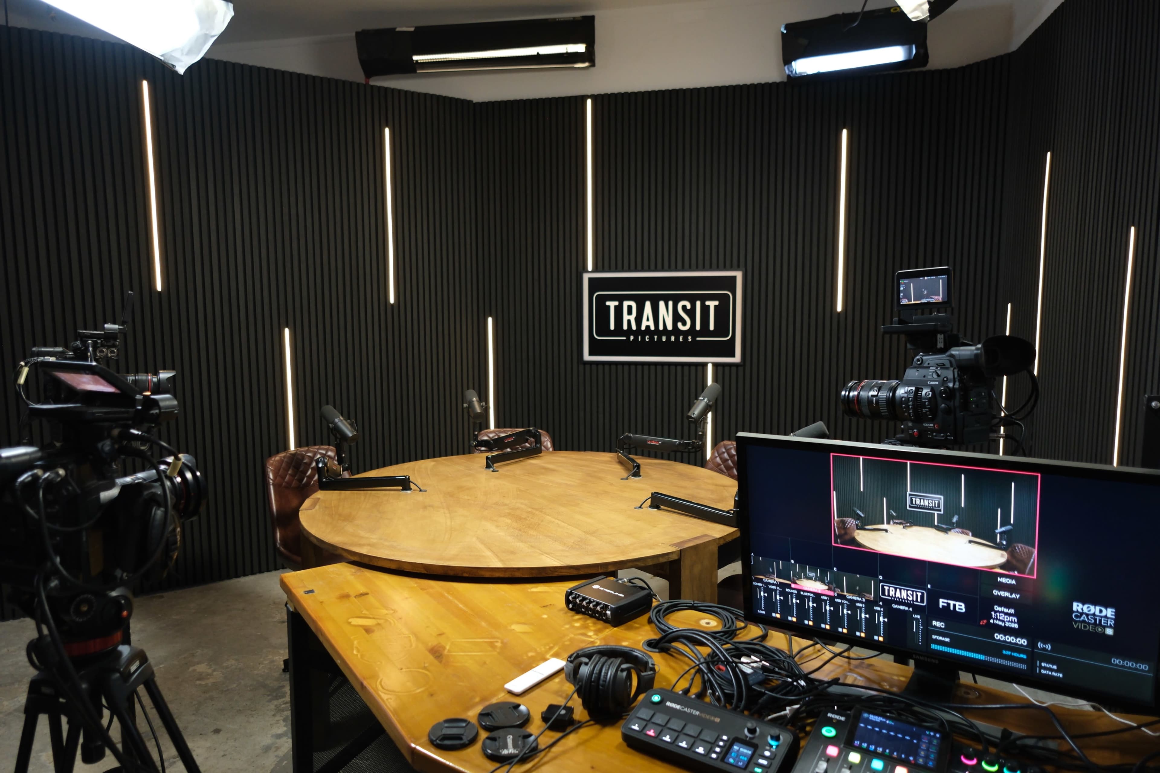 The image shows a modern recording studio with a circular wooden table, multiple cameras, and a control panel, featuring a backdrop with the word "TRANSIT."