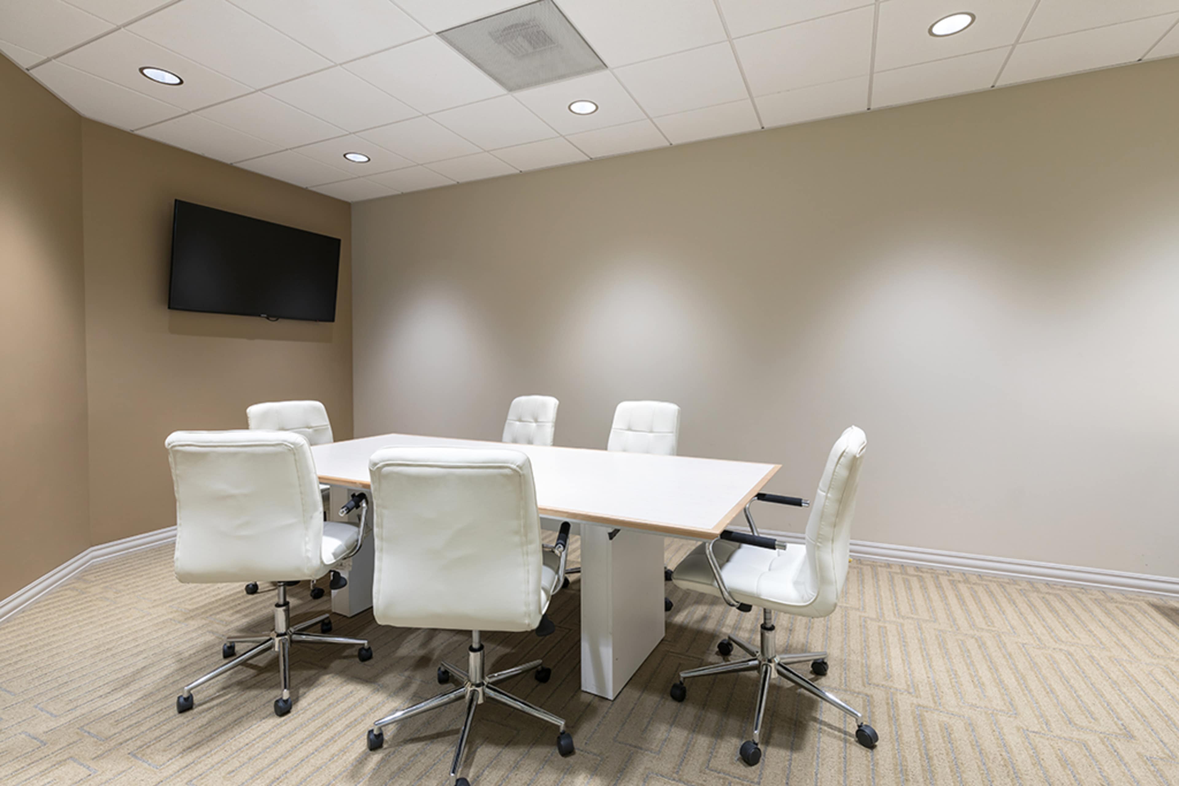 A modern conference room features a large white table surrounded by five white chairs and a wall-mounted television.