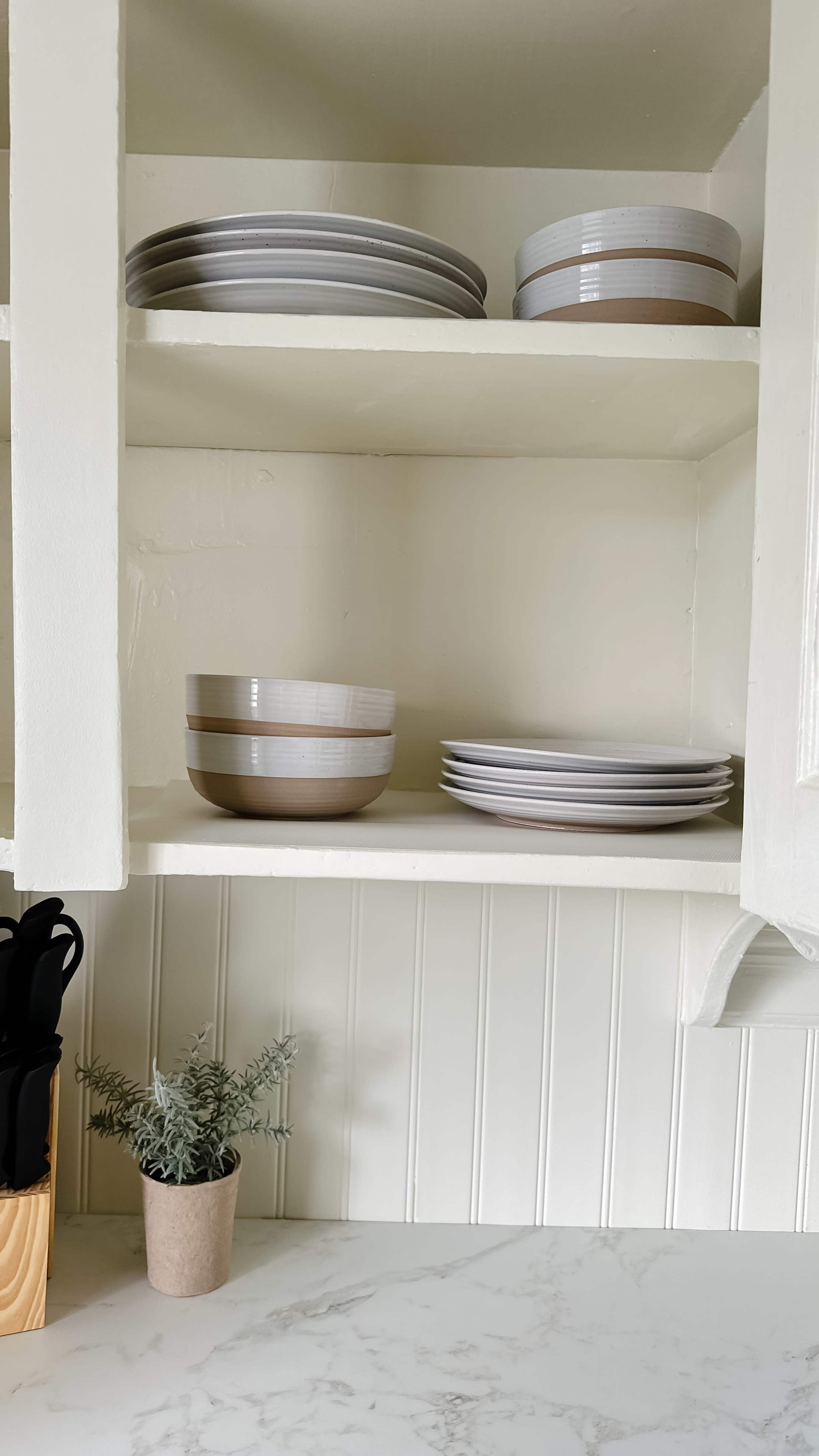 The image shows neatly arranged bowls and plates in a white kitchen cabinet, with a small potted plant on the shelf below.