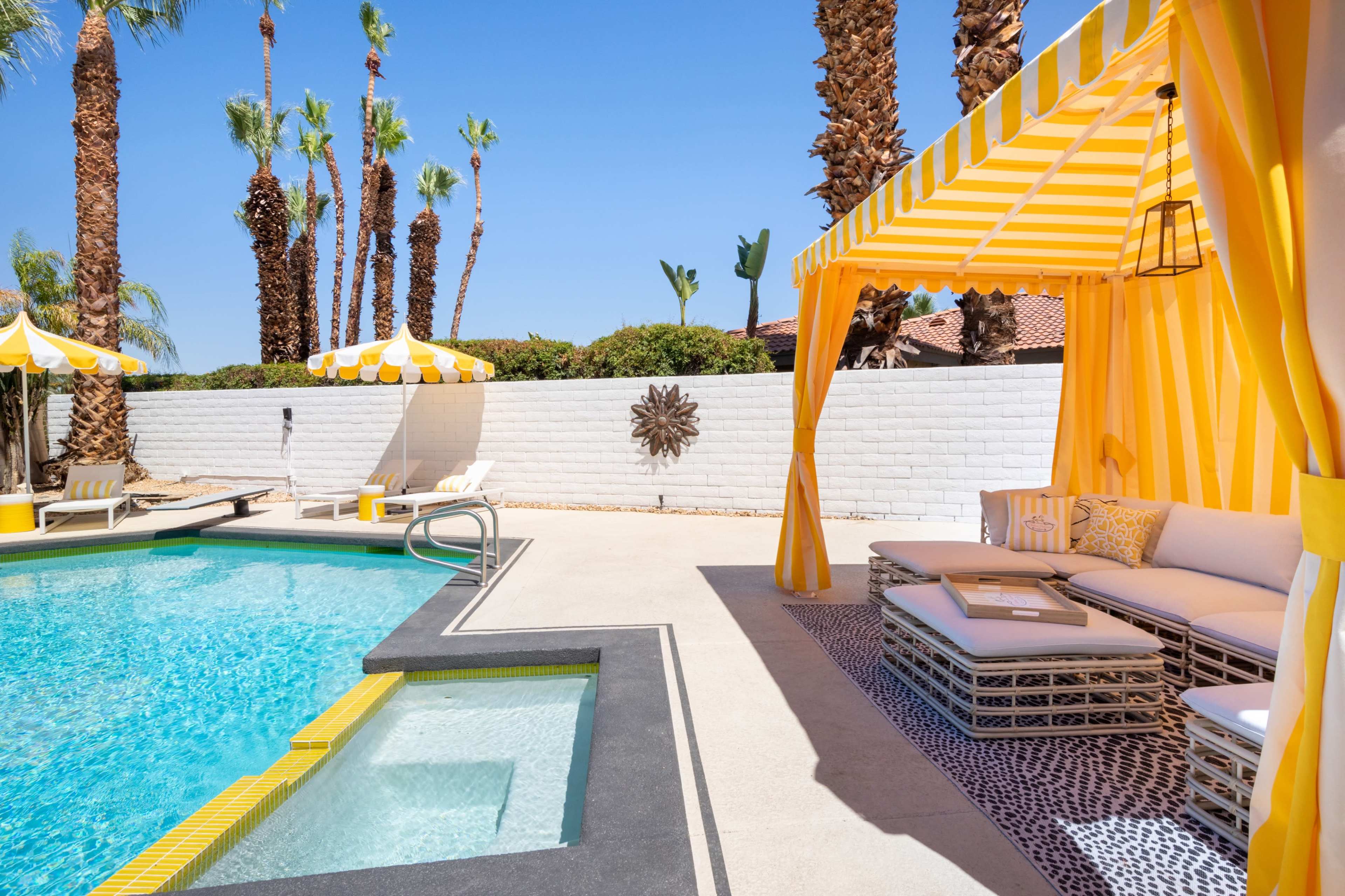 The scene features a swimming pool surrounded by tropical palm trees, a sunbathing area with yellow-striped umbrellas, and a shaded seating area with a couch.