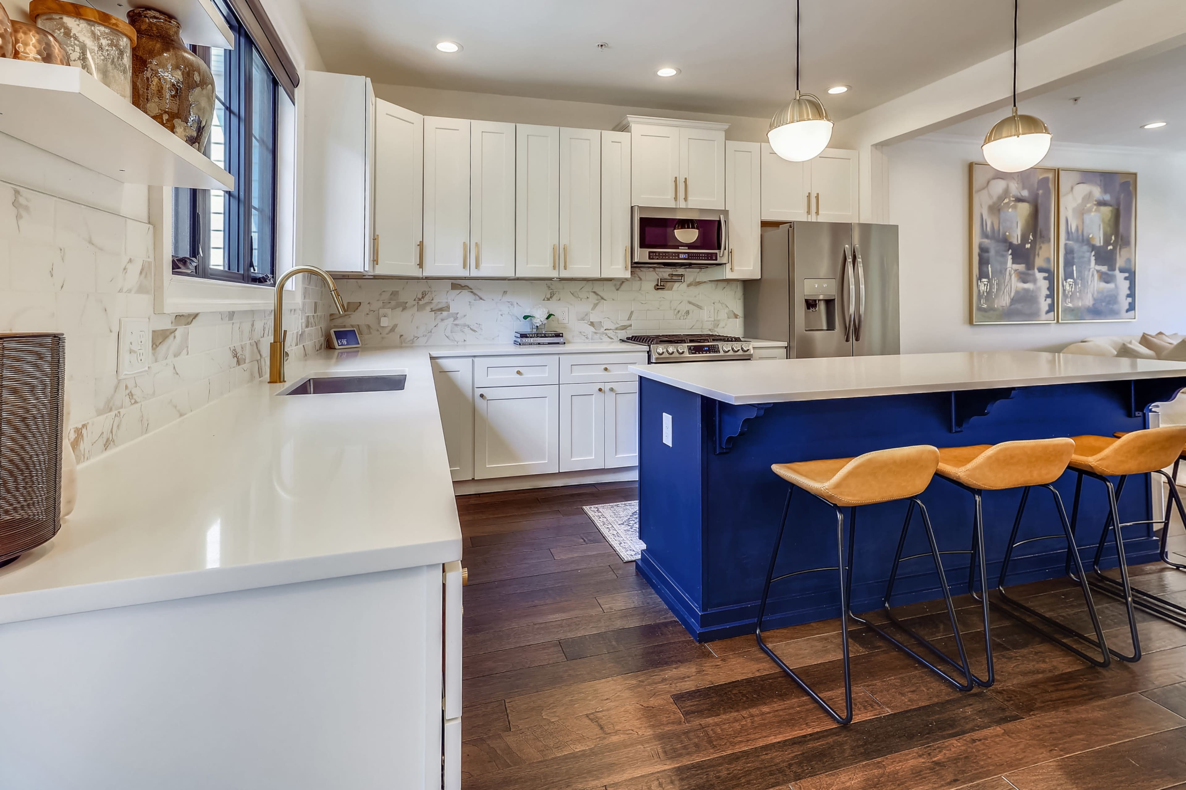 A modern kitchen with white cabinets, a marble backsplash, an island with blue cabinetry, and three bar stools.