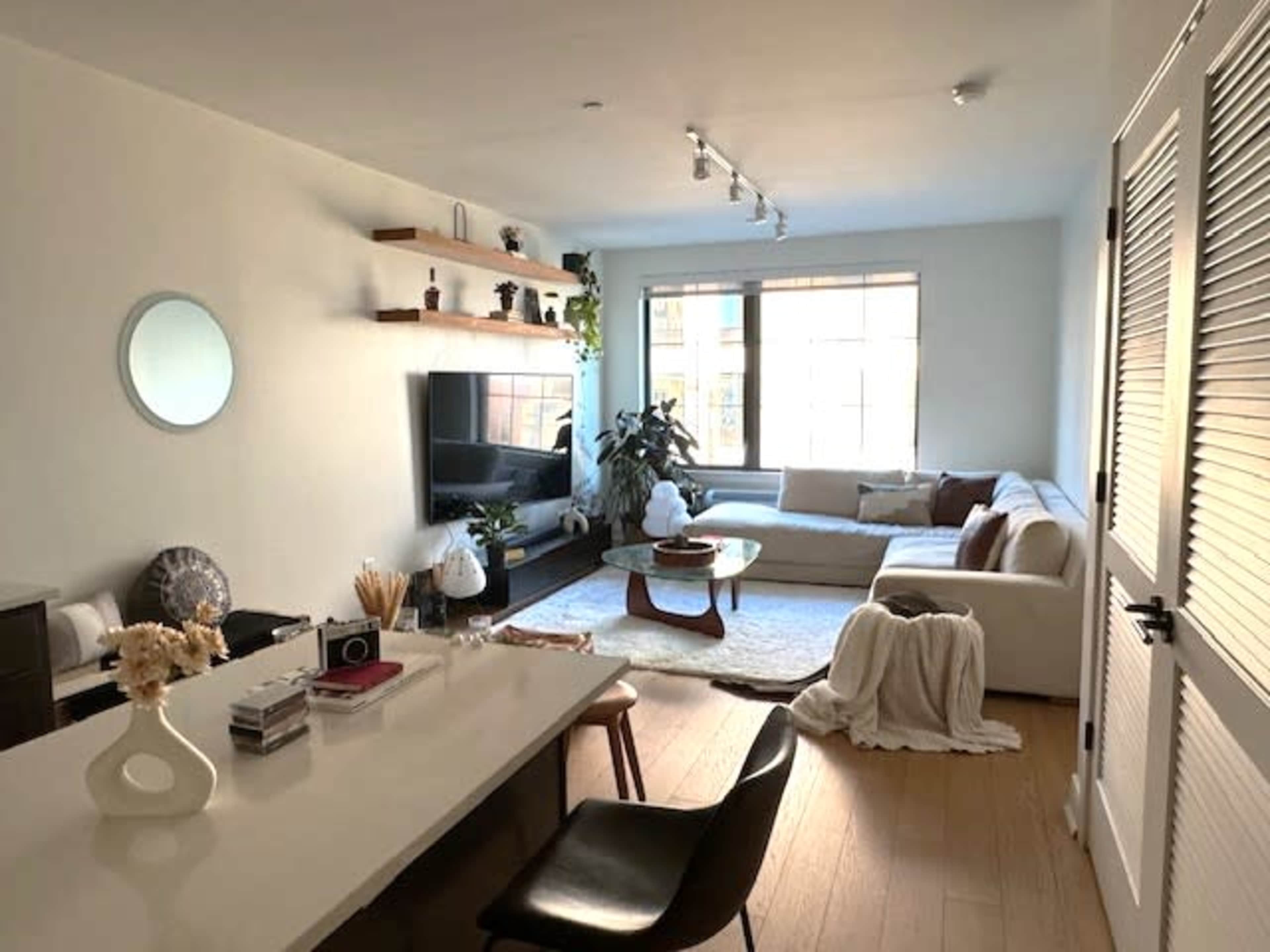 A modern living room with a sectional sofa, a coffee table, and a television mounted on the wall, alongside a kitchen area with sleek cabinetry and minimalist decor.