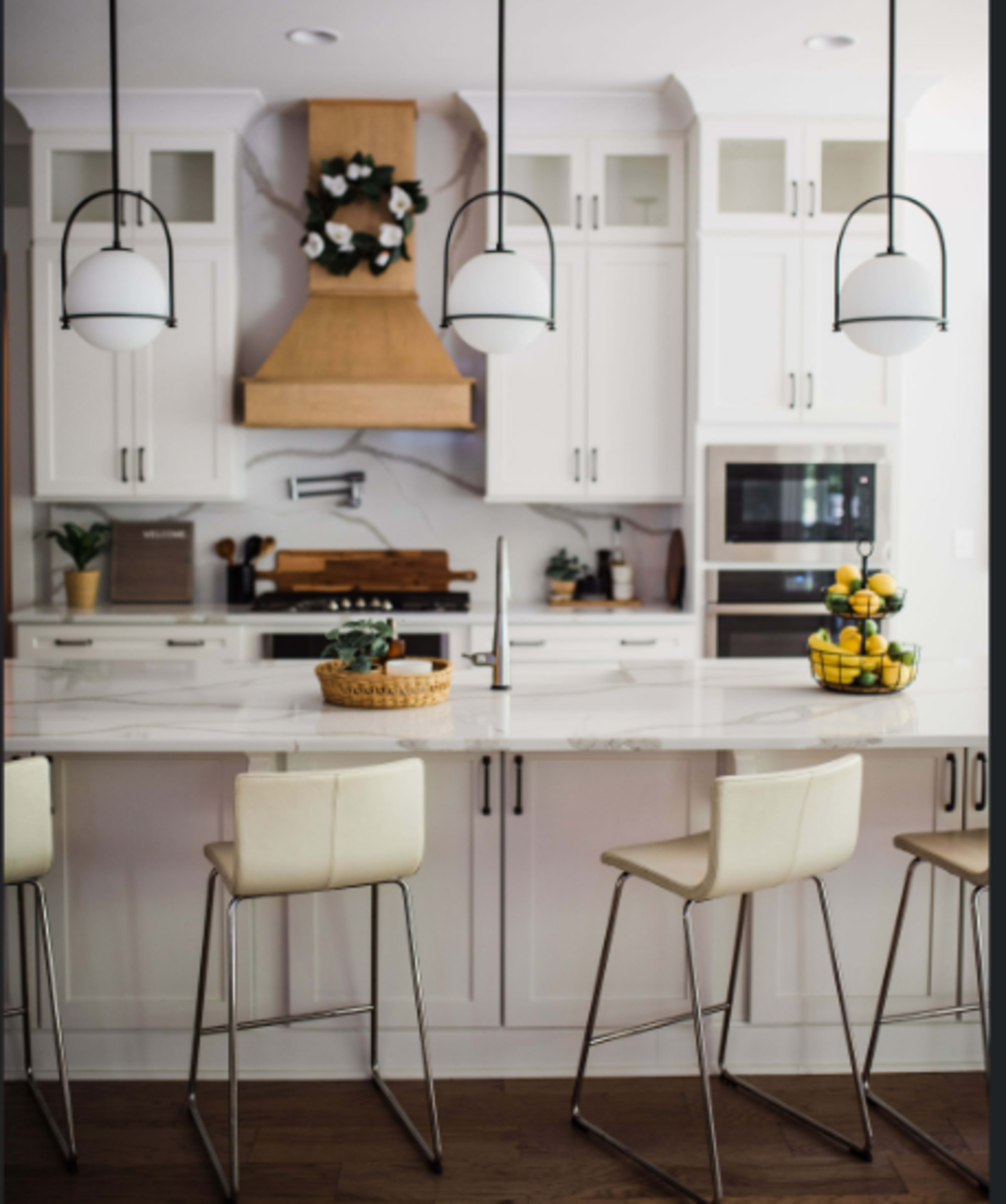 A modern kitchen features white cabinetry, a large marble island with four beige stools, and a wooden range hood above the stove.