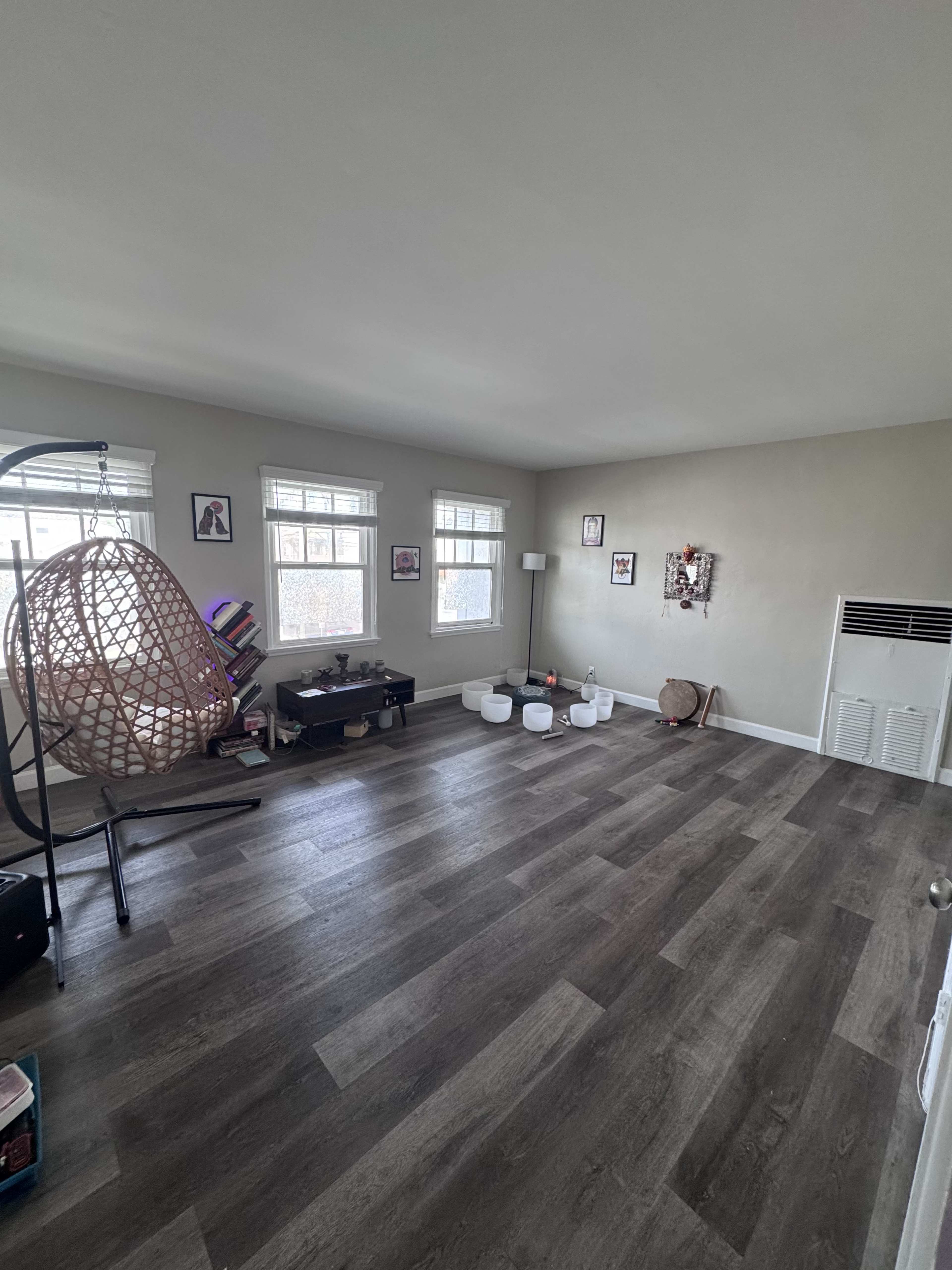 The image shows a spacious room with modern flooring, featuring a hanging chair, a low table, and several decorative items against light-colored walls with windows letting in natural light.