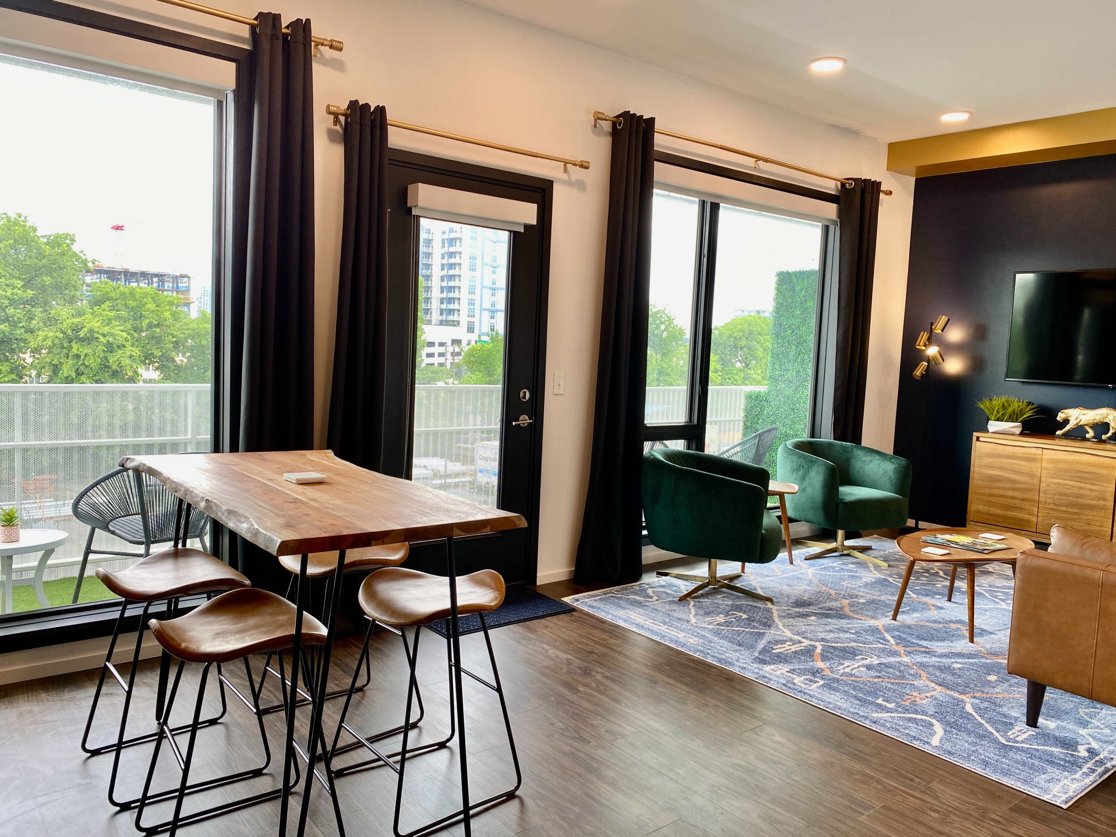 A modern apartment living area featuring a wooden dining table with stools, two green accent chairs, a black door leading to a balcony, and large windows letting in natural light.