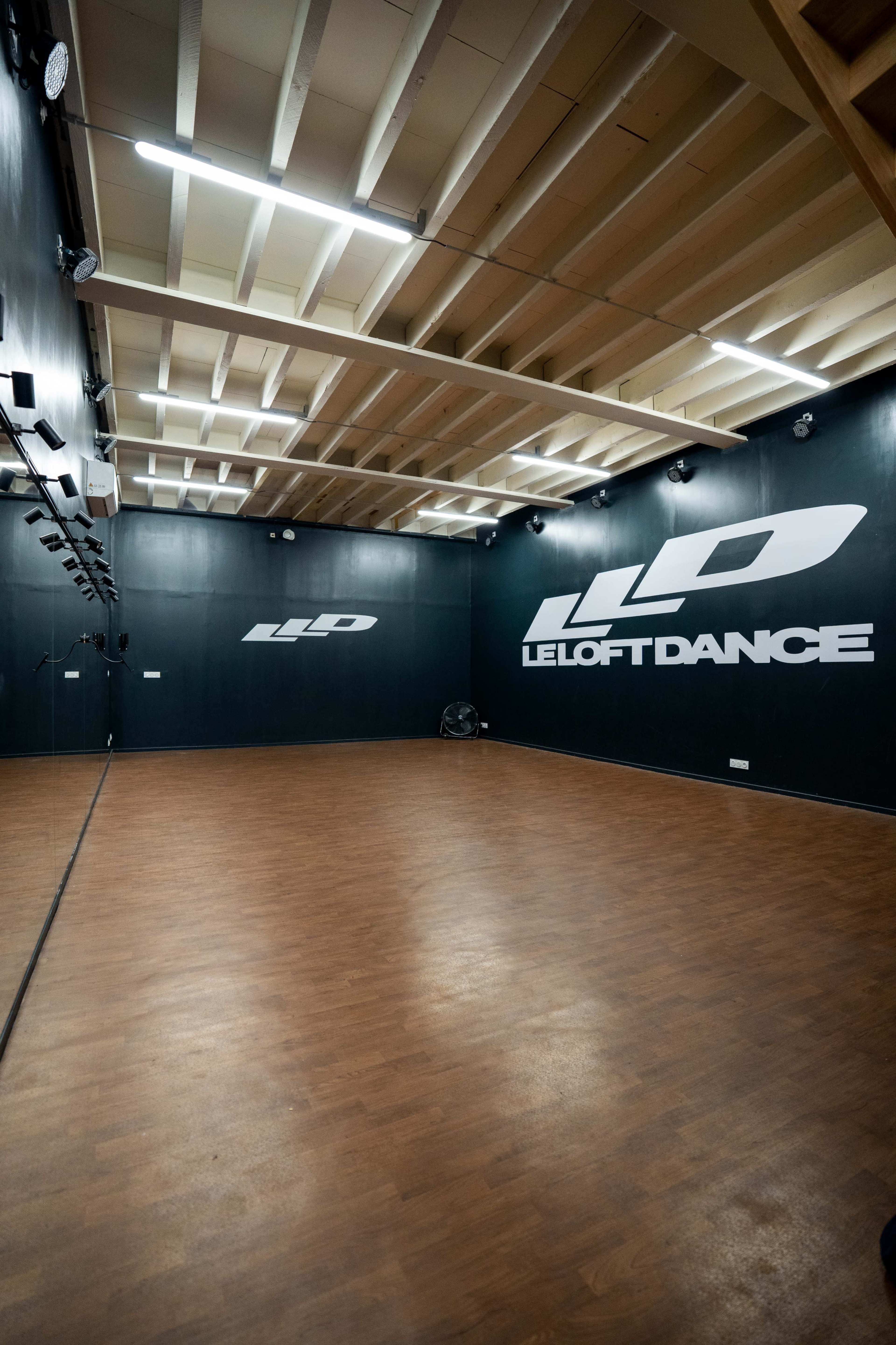 The image shows an empty dance studio with dark walls, wooden flooring, and a logo on the wall.