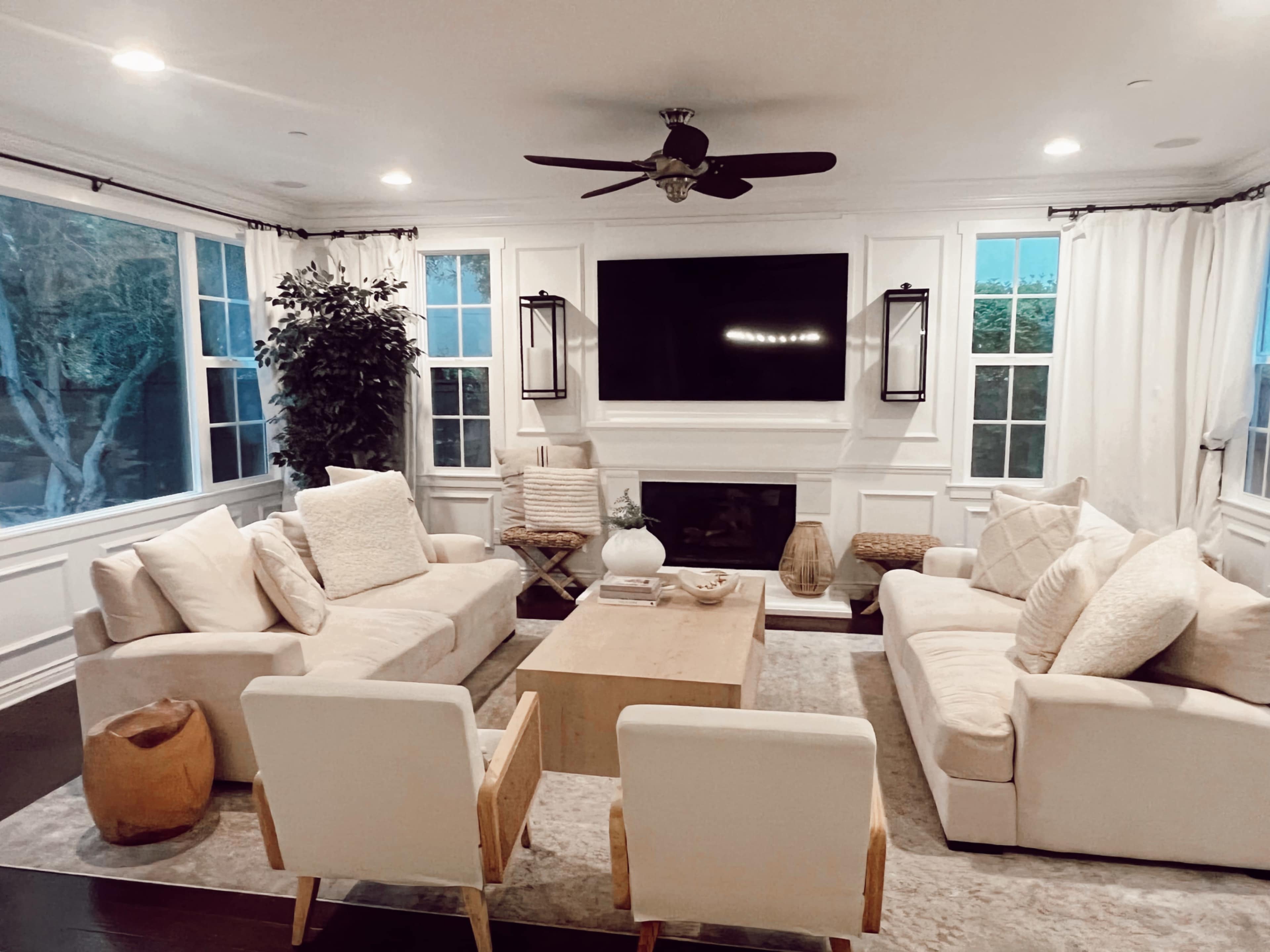 A modern living room features two beige sofas and two armchairs arranged around a central wooden coffee table, with a large flat-screen TV mounted on the wall above a white mantel.
