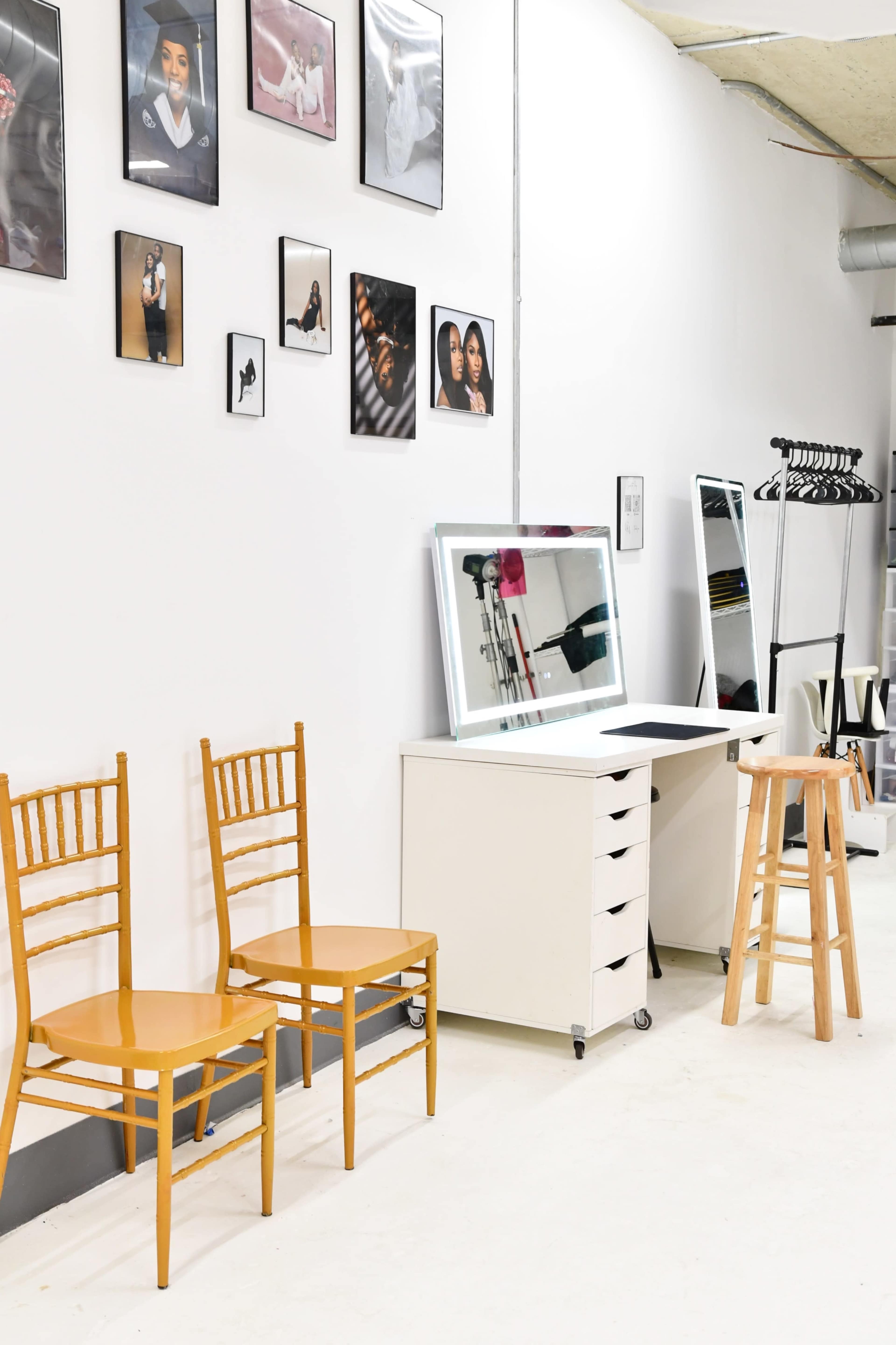 The image shows a bright, minimalistic room with wooden chairs, a white table and storage unit, a mirror, and fashion photographs displayed on the wall.