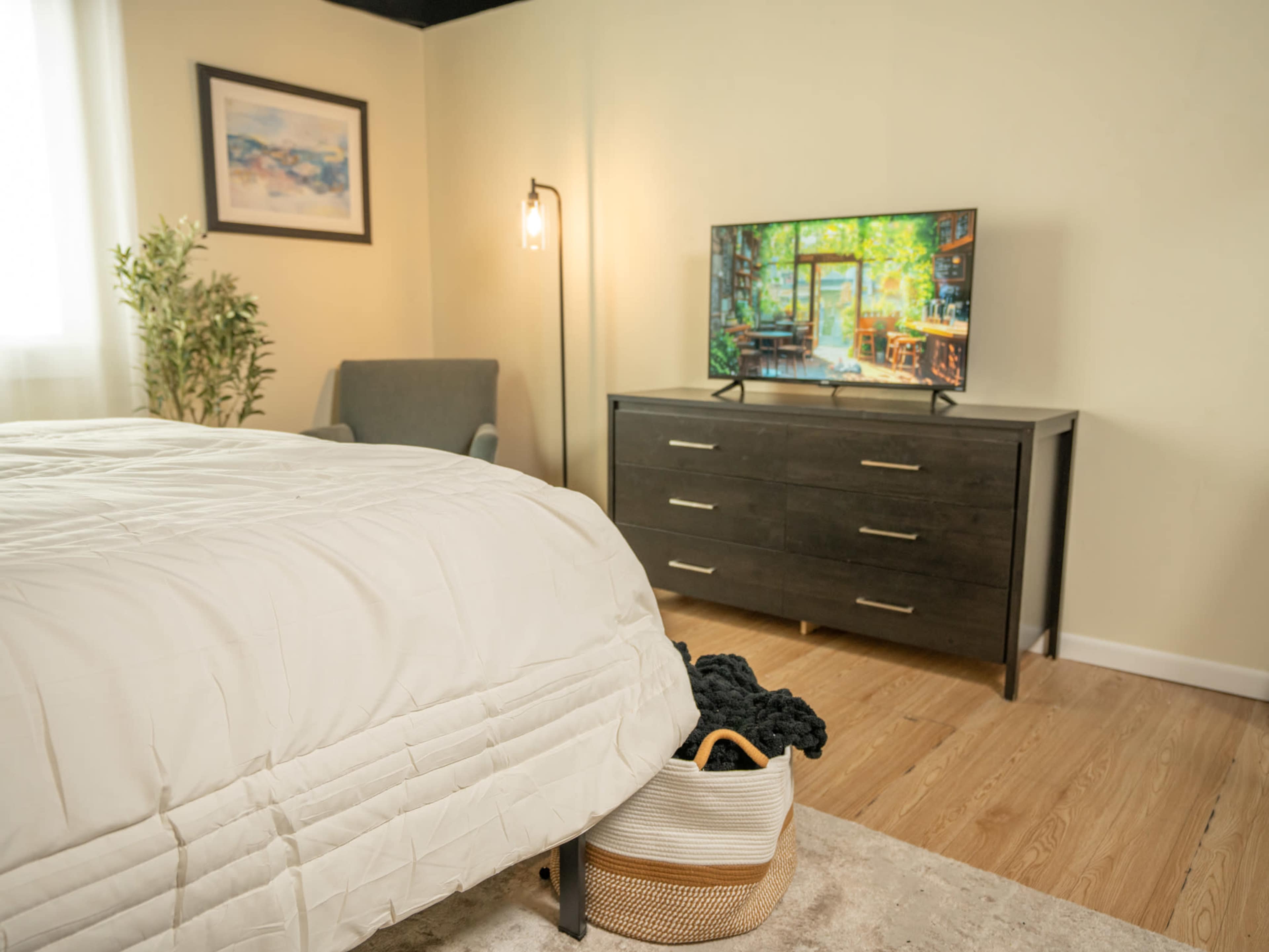 A well-furnished bedroom features a bed with a light comforter, a gray armchair, a dresser, and a television displaying a garden scene.