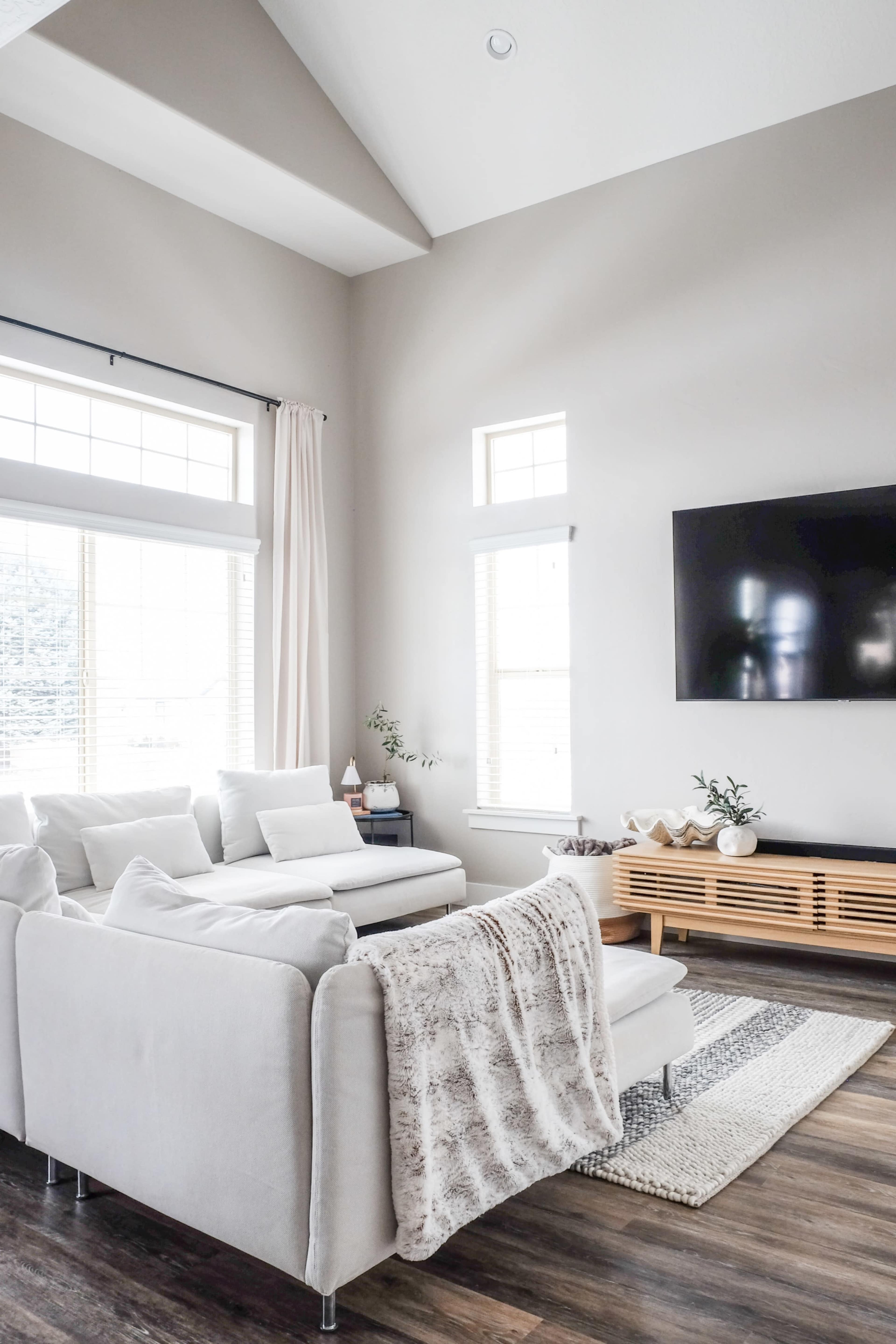 A modern living room features a light-colored sectional sofa, a wooden TV stand, large windows with sheer curtains, and a rug on the hardwood floor.