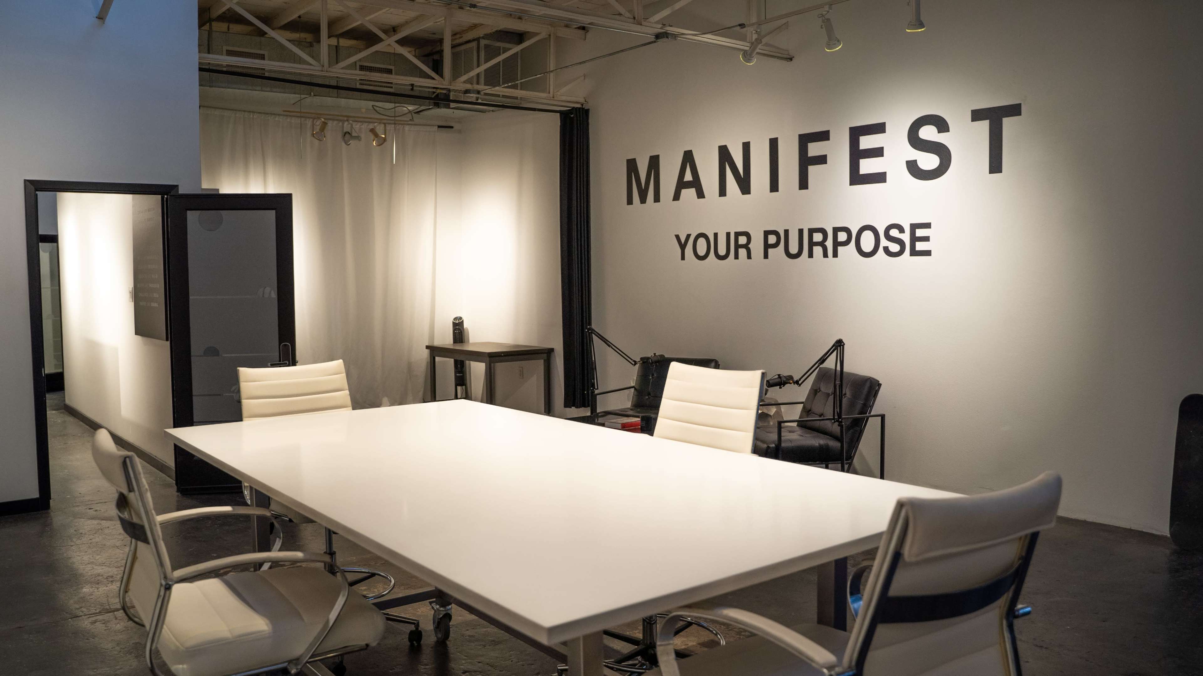 A large, minimalist conference room features a white table, several chairs, and a wall with the words "MANIFEST YOUR PURPOSE" displayed prominently.