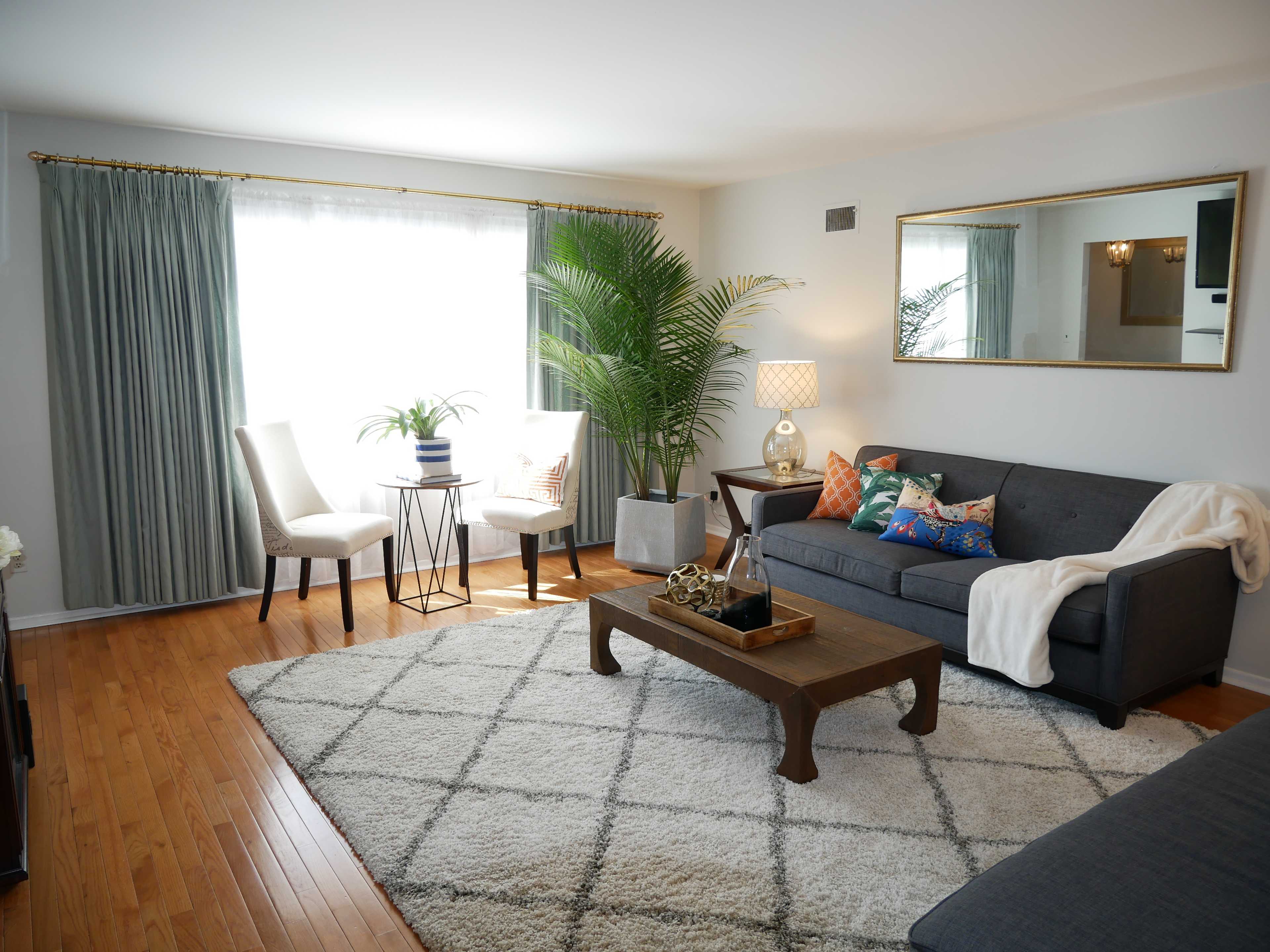 The living room features a gray sofa, a wooden coffee table, two chairs, and a potted plant, all set on a patterned rug with large windows allowing natural light.