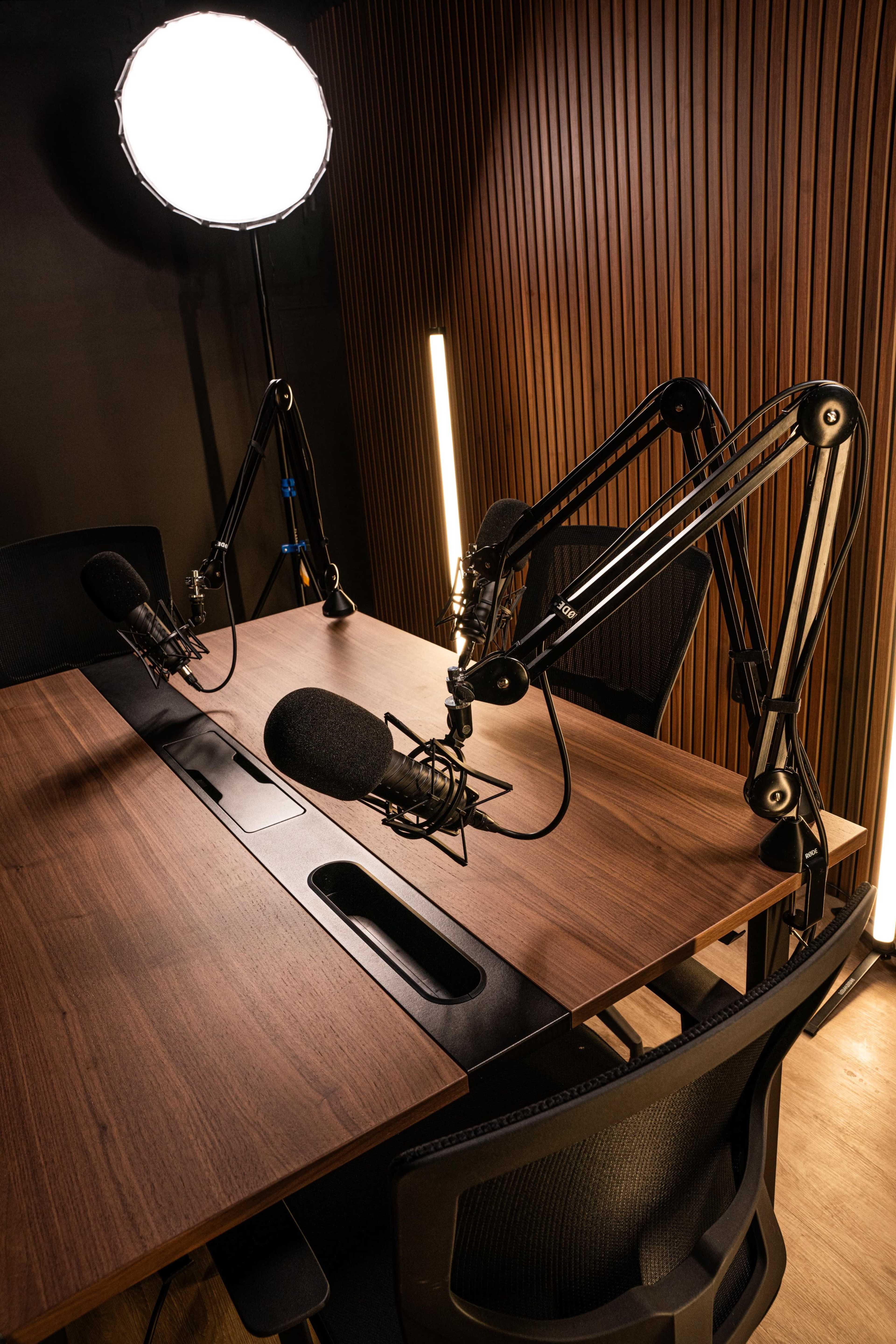The image shows a podcast recording studio with two microphones mounted on adjustable arms, a wooden table, and professional lighting.