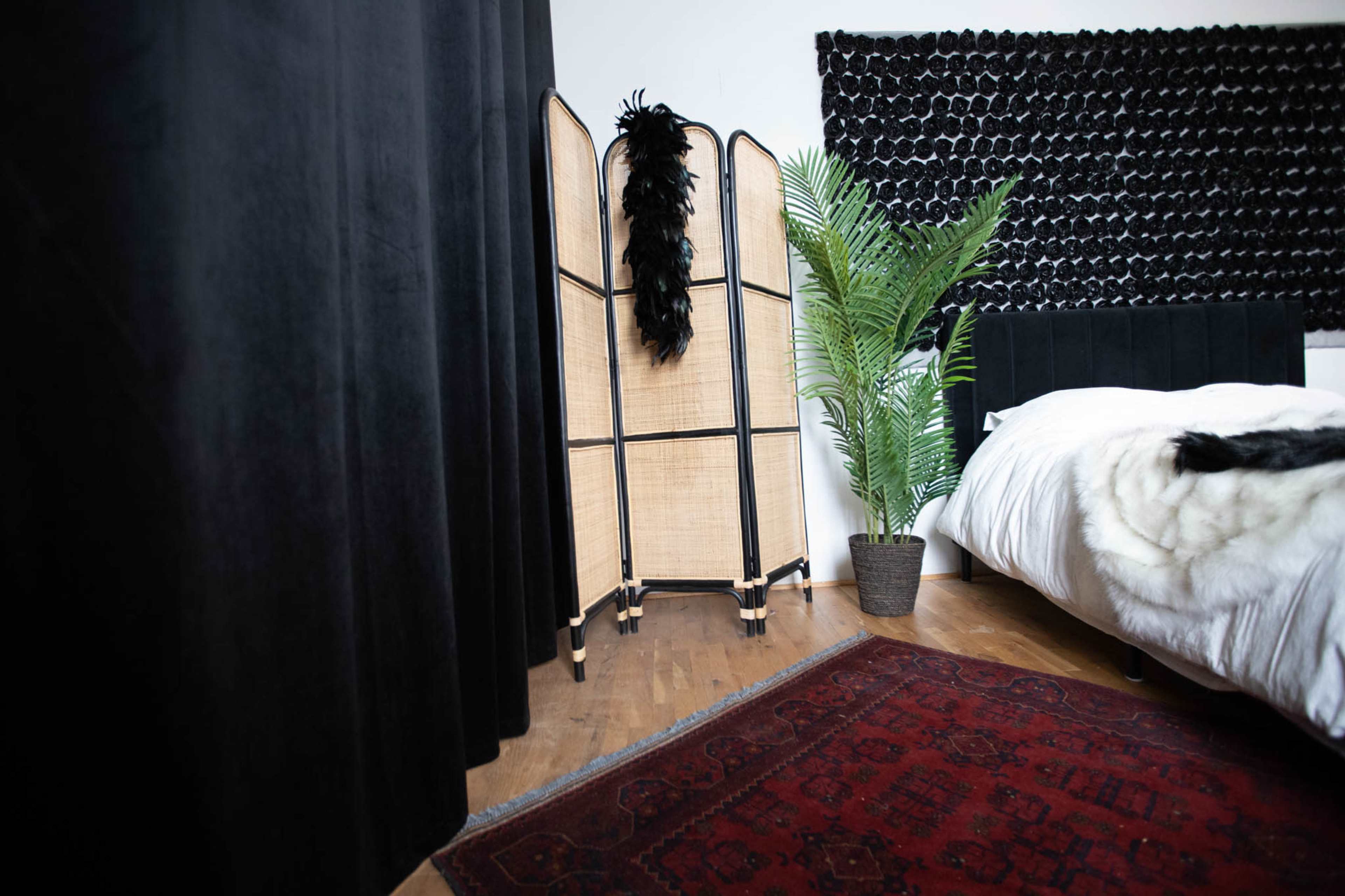 A bedroom corner featuring a folding screen, a potted plant, and a bed with a white duvet against a textured black wall.