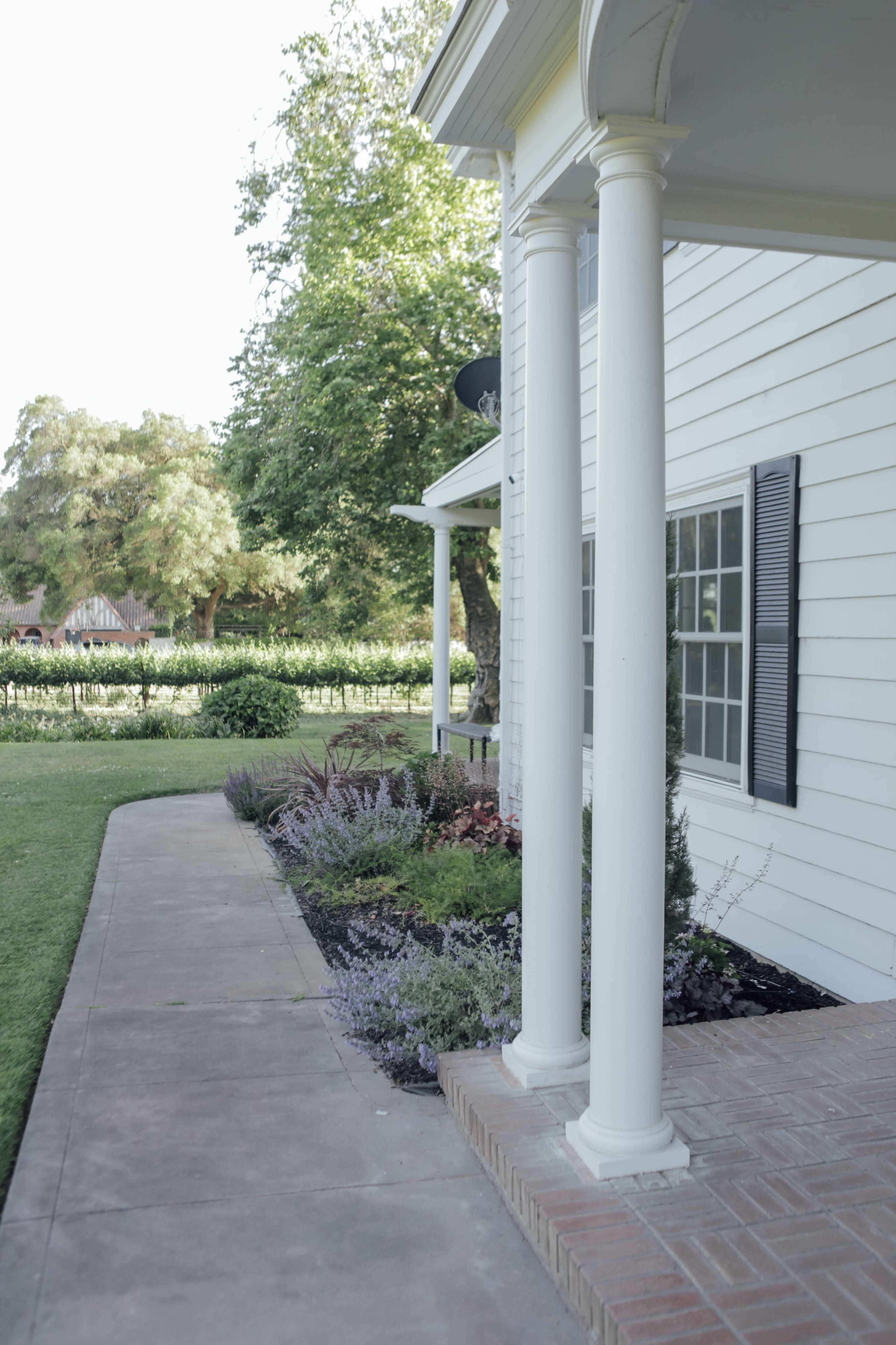 The image shows a white house with two columns on a porch, surrounded by a landscaped garden along a concrete pathway.
