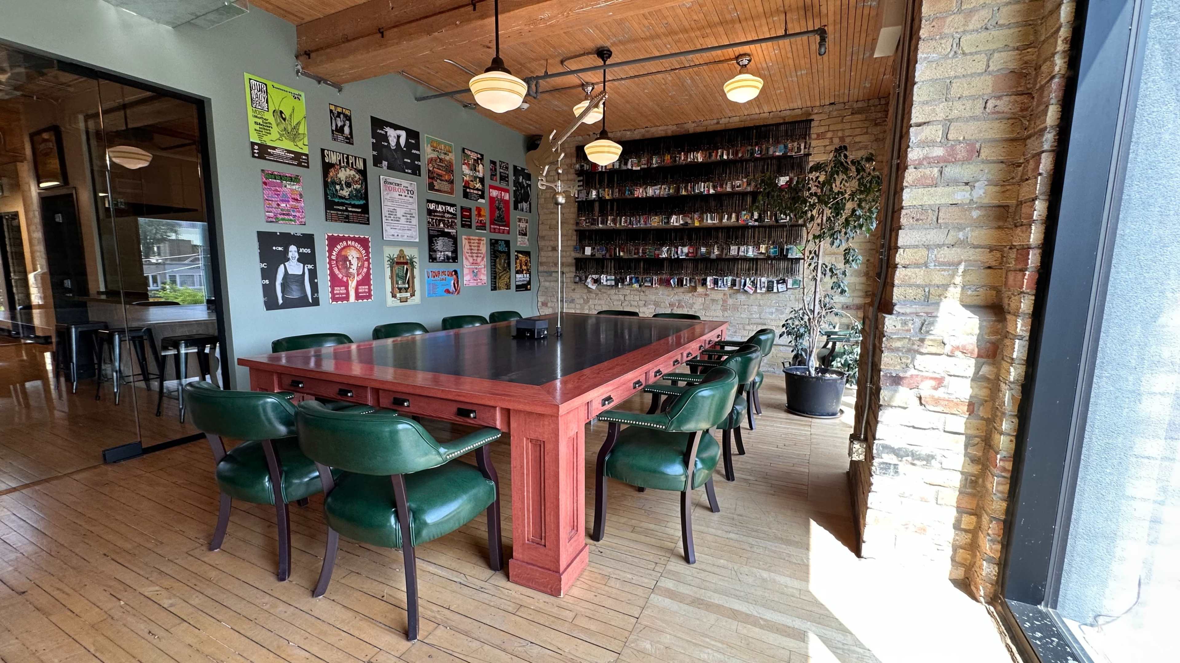 The image shows a spacious dining area featuring a large wooden table surrounded by green chairs, with brick walls adorned with posters and a wall of shelves in the background.