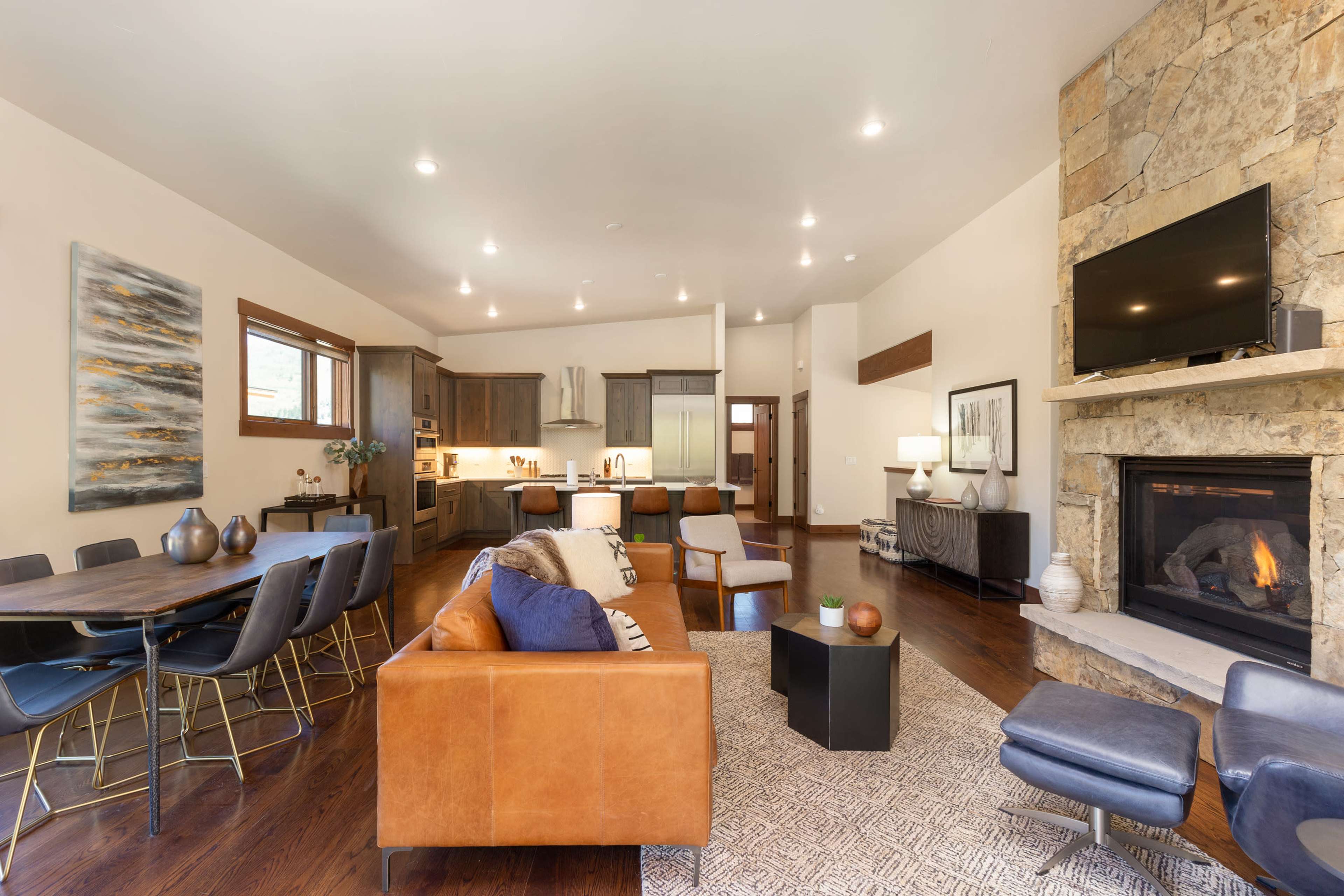 A modern living area features a stone fireplace, sectional sofa, dining table, and kitchen in an open layout with wooden floors.