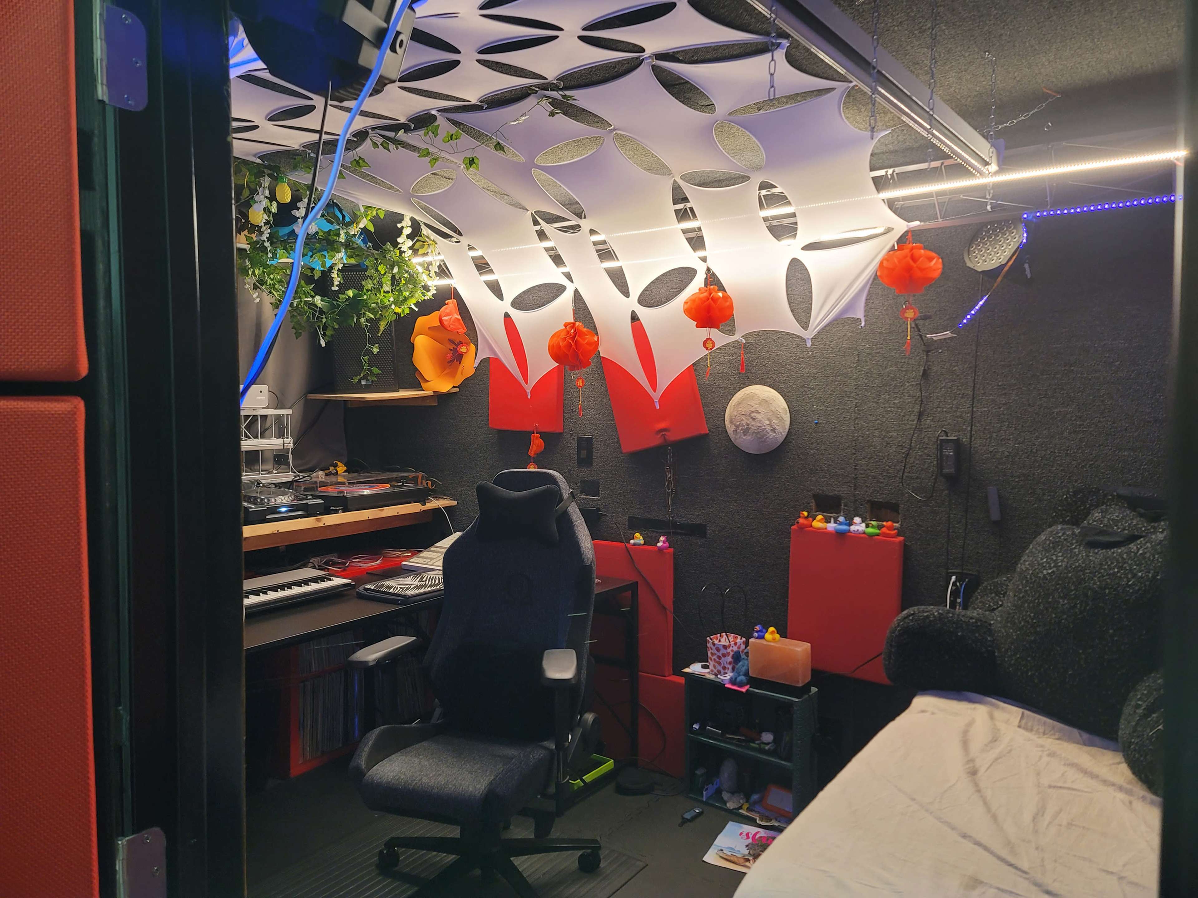 A small music production studio with a black chair, sound equipment, decorative plants, and colorful hanging ornaments.
