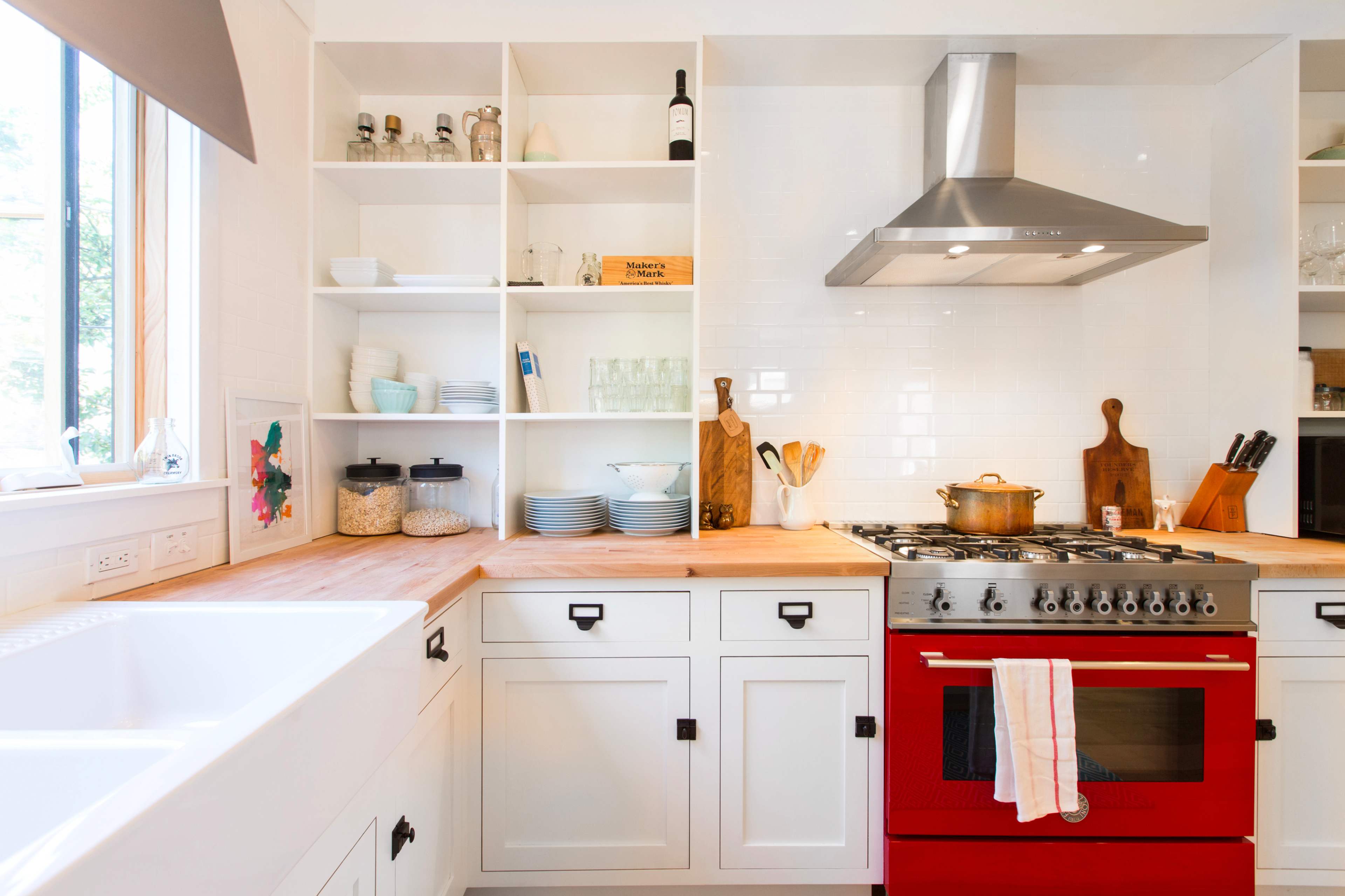 A modern kitchen features a red stove, wooden countertops, open shelving with dishes and jars, and a large sink by a window.