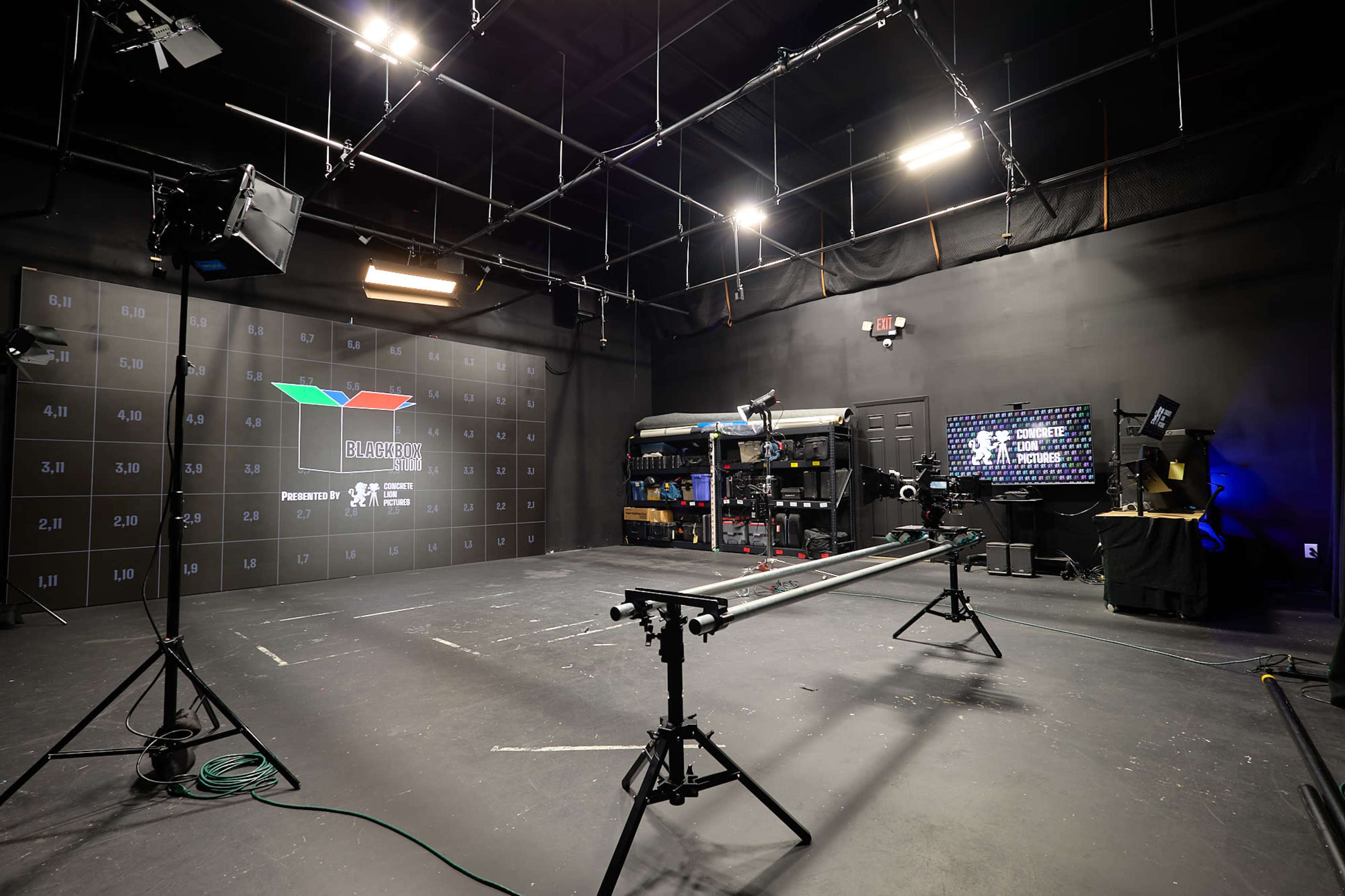 The image shows a dark studio space equipped with camera tracks, lighting setups, and a backdrop featuring a graphic display on the wall.