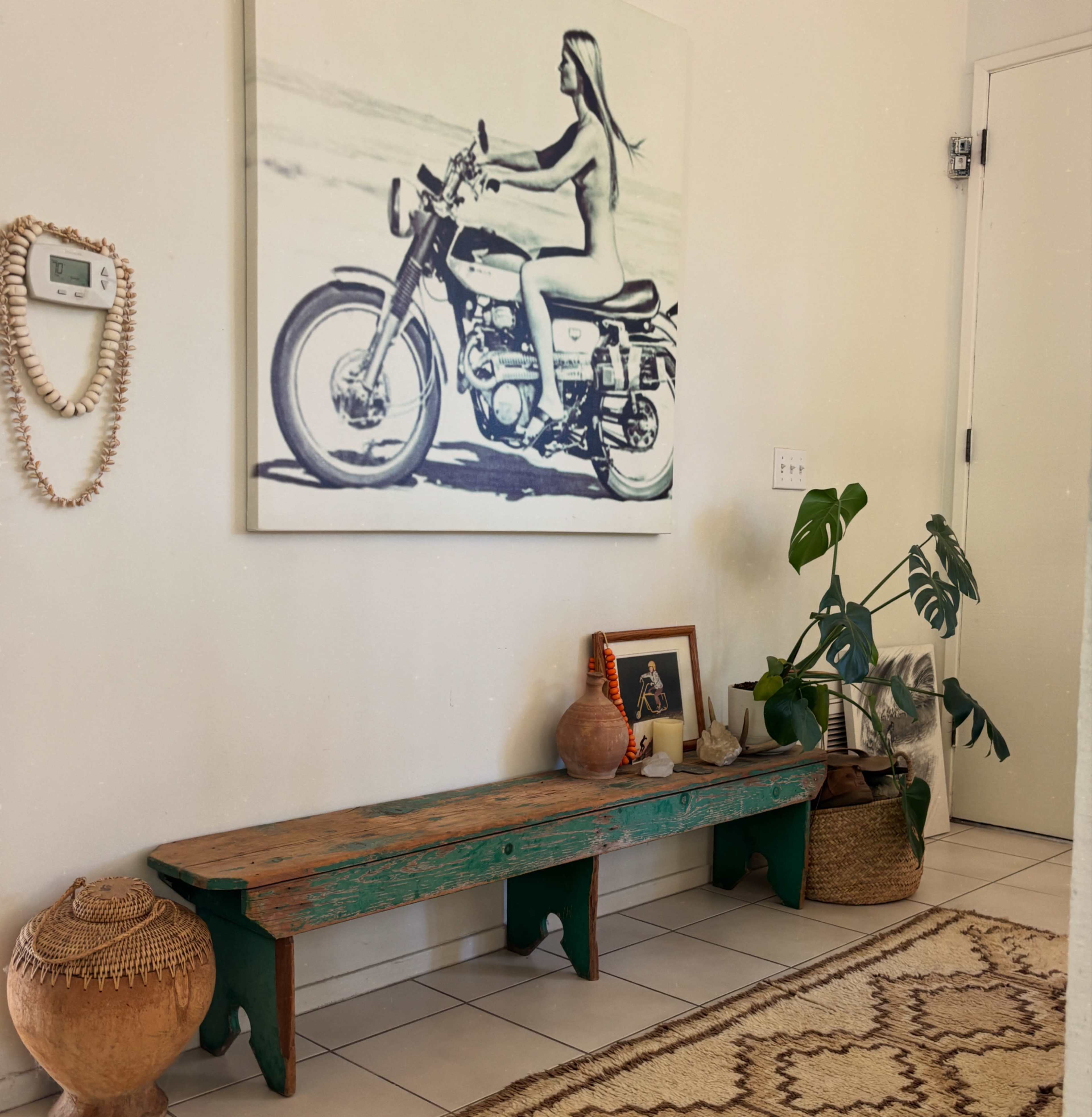The image shows an interior space featuring a wooden bench, a large wall art of a woman on a motorcycle, and various decorative objects including pots and a plant near the wall.