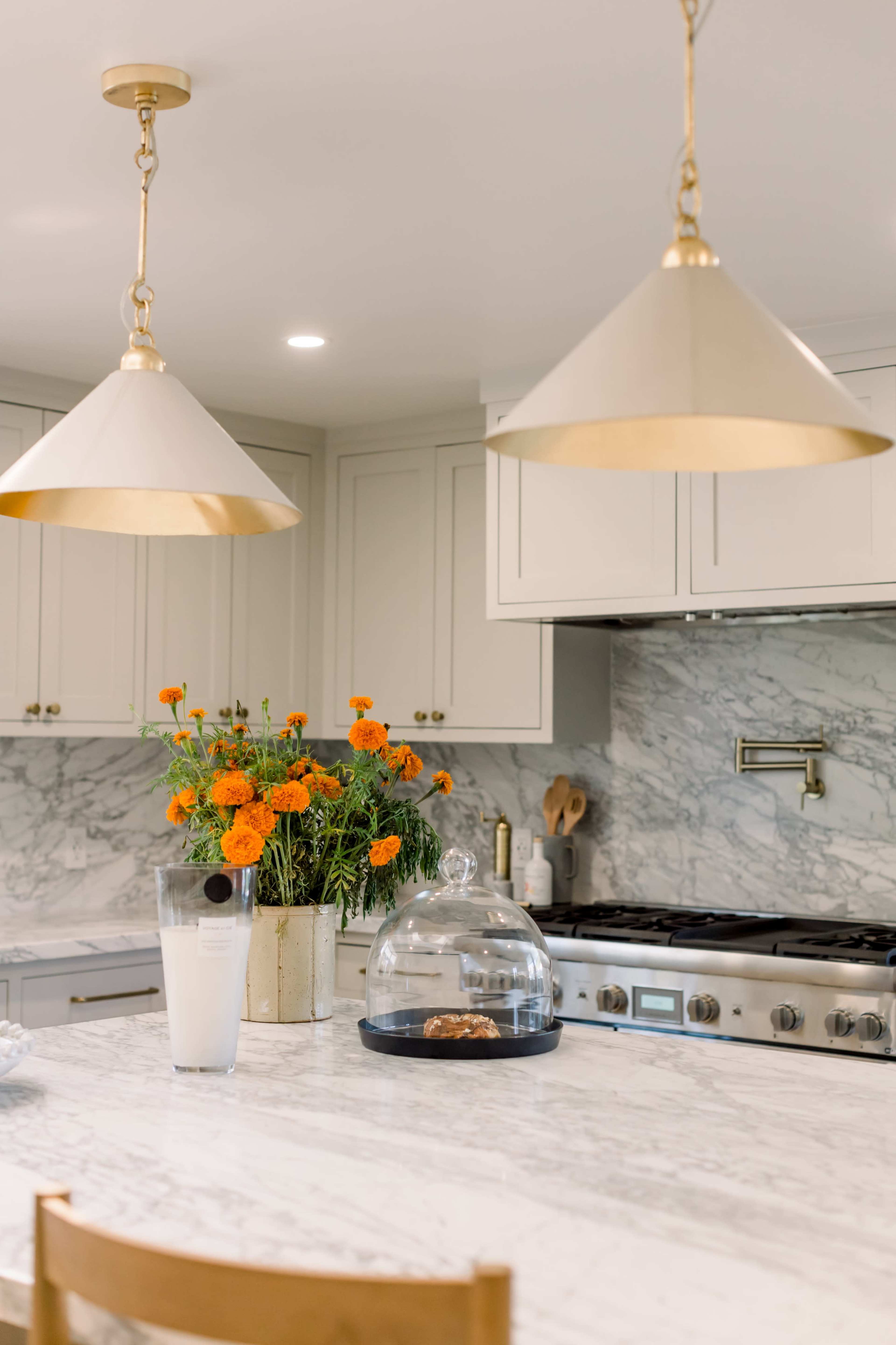 The kitchen features a marble countertop with a glass cake dome, under two gold pendant lights, and a vase of orange marigolds on a nearby surface.