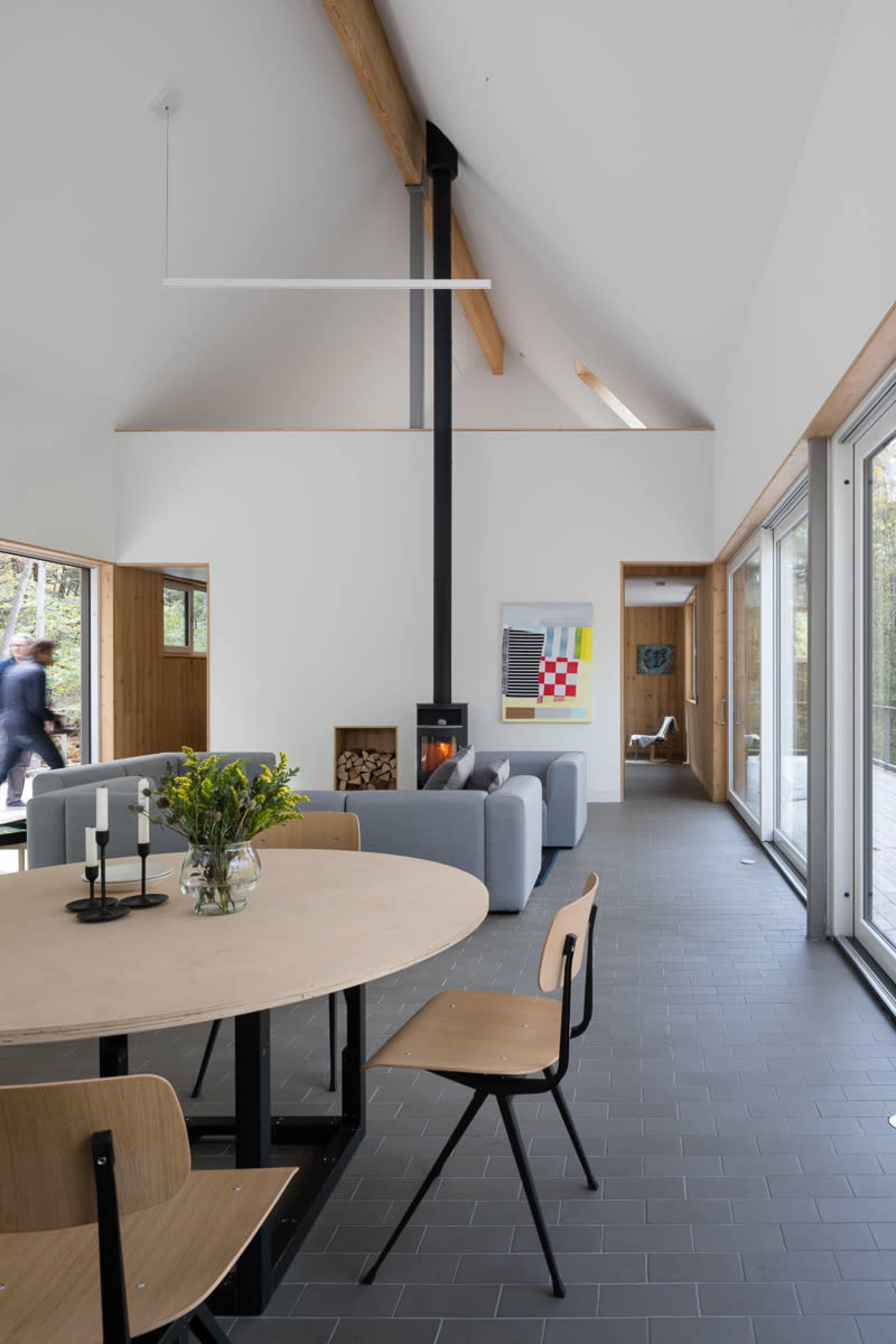 A modern interior features a dining table with black metal legs, surrounded by wooden chairs, and a living area with a fireplace and large glass doors opening to an outdoor space.