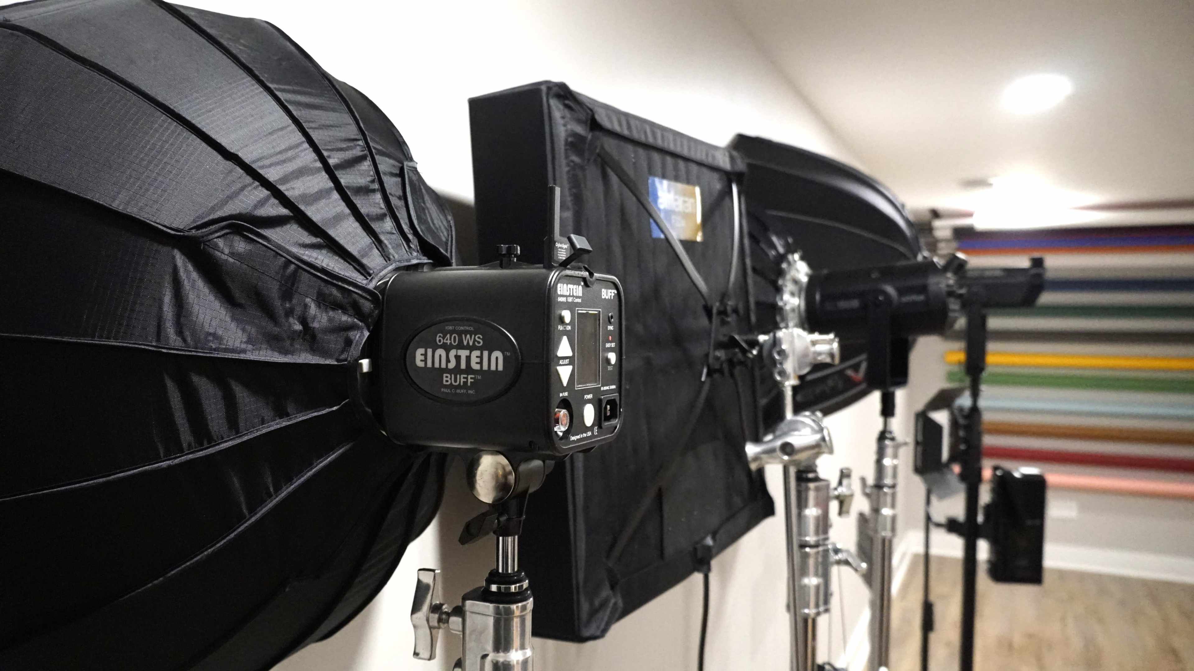 The image shows studio lighting equipment with softboxes and reflectors mounted on stands against a wall with colorful stripes.