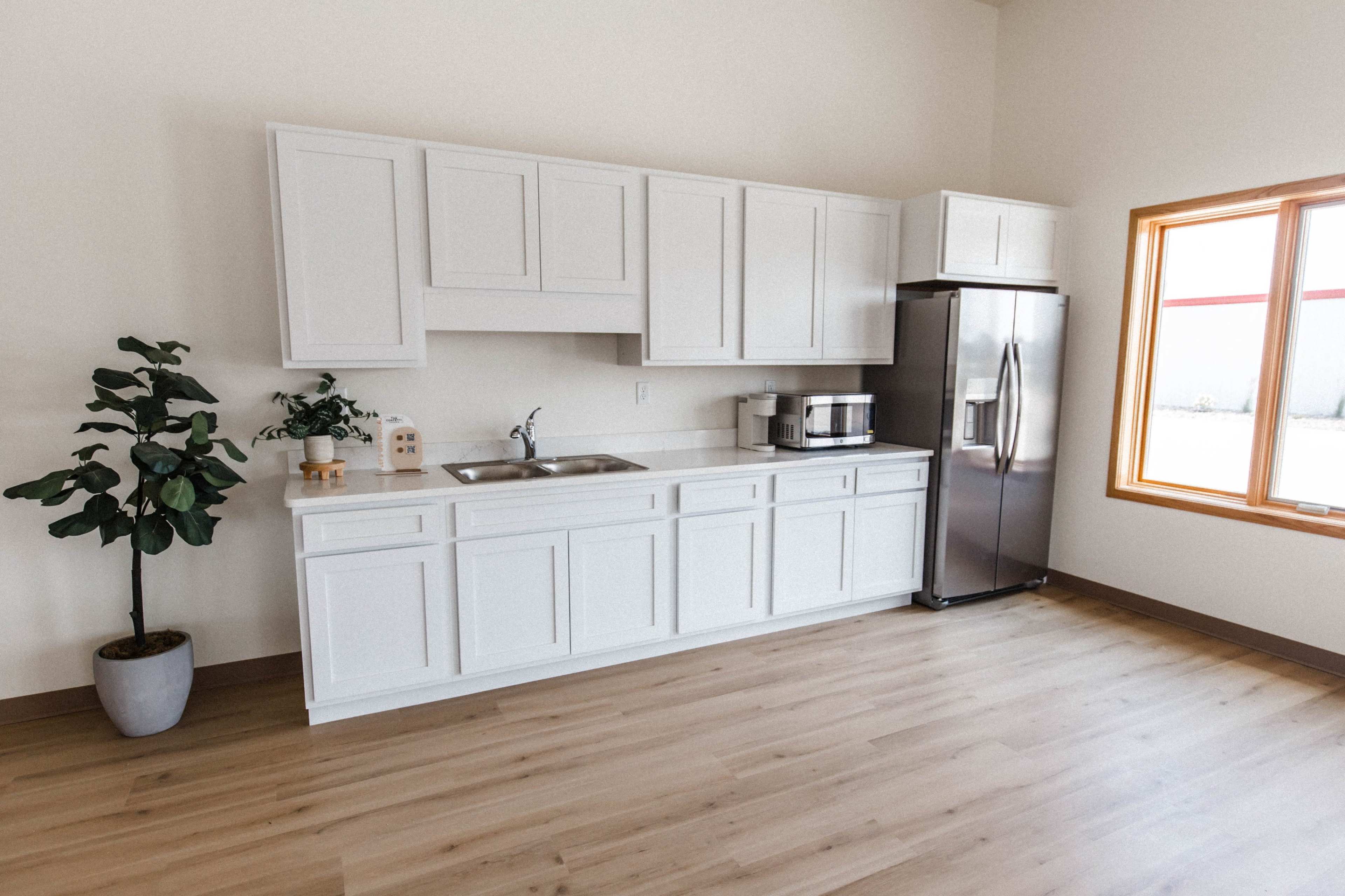 A modern kitchen with white cabinetry, a stainless steel refrigerator, and a sink, illuminated by natural light from a large window.
