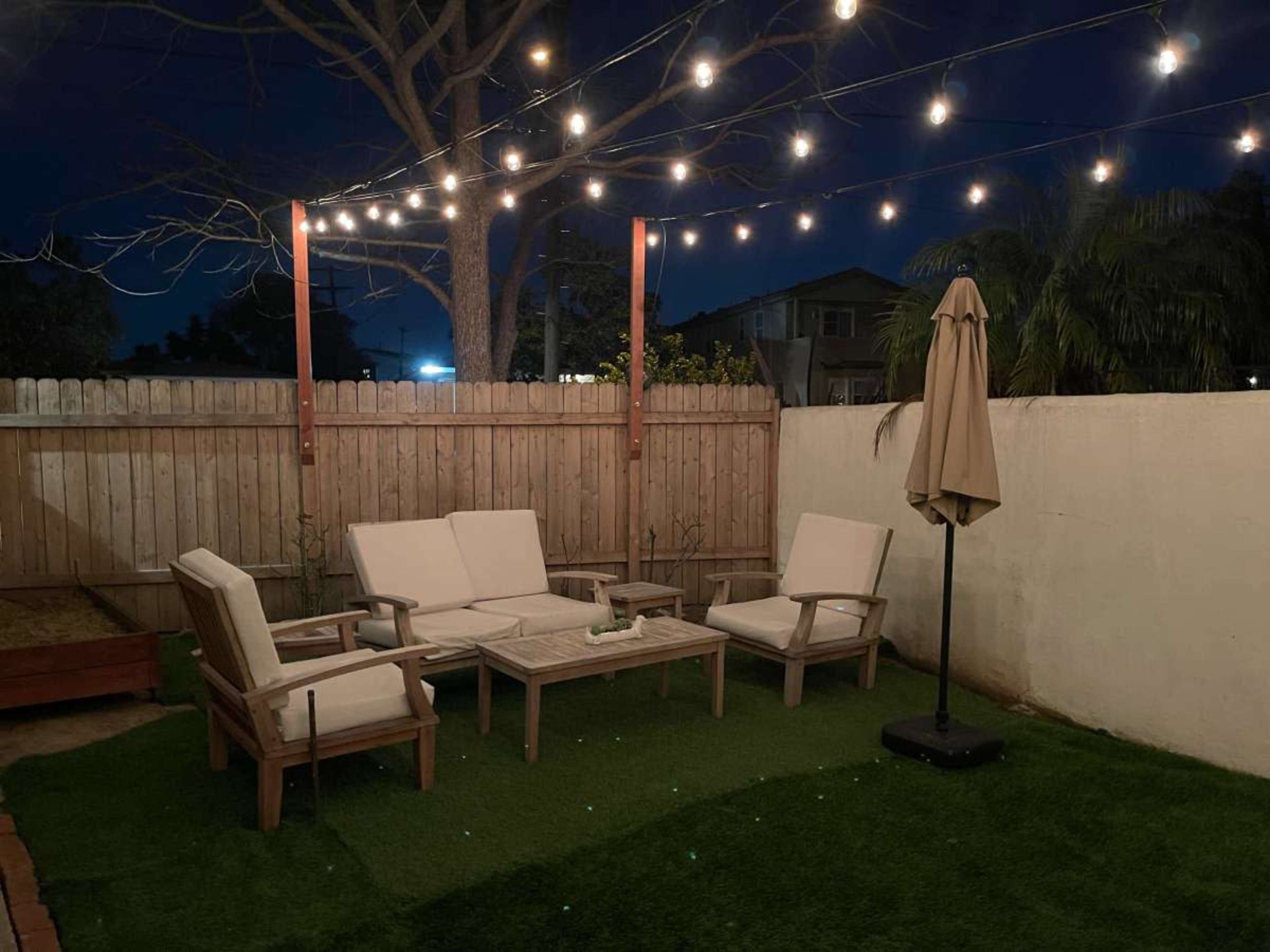 A cozy outdoor seating area features wooden furniture and string lights overhead, set against a fenced backyard at night.