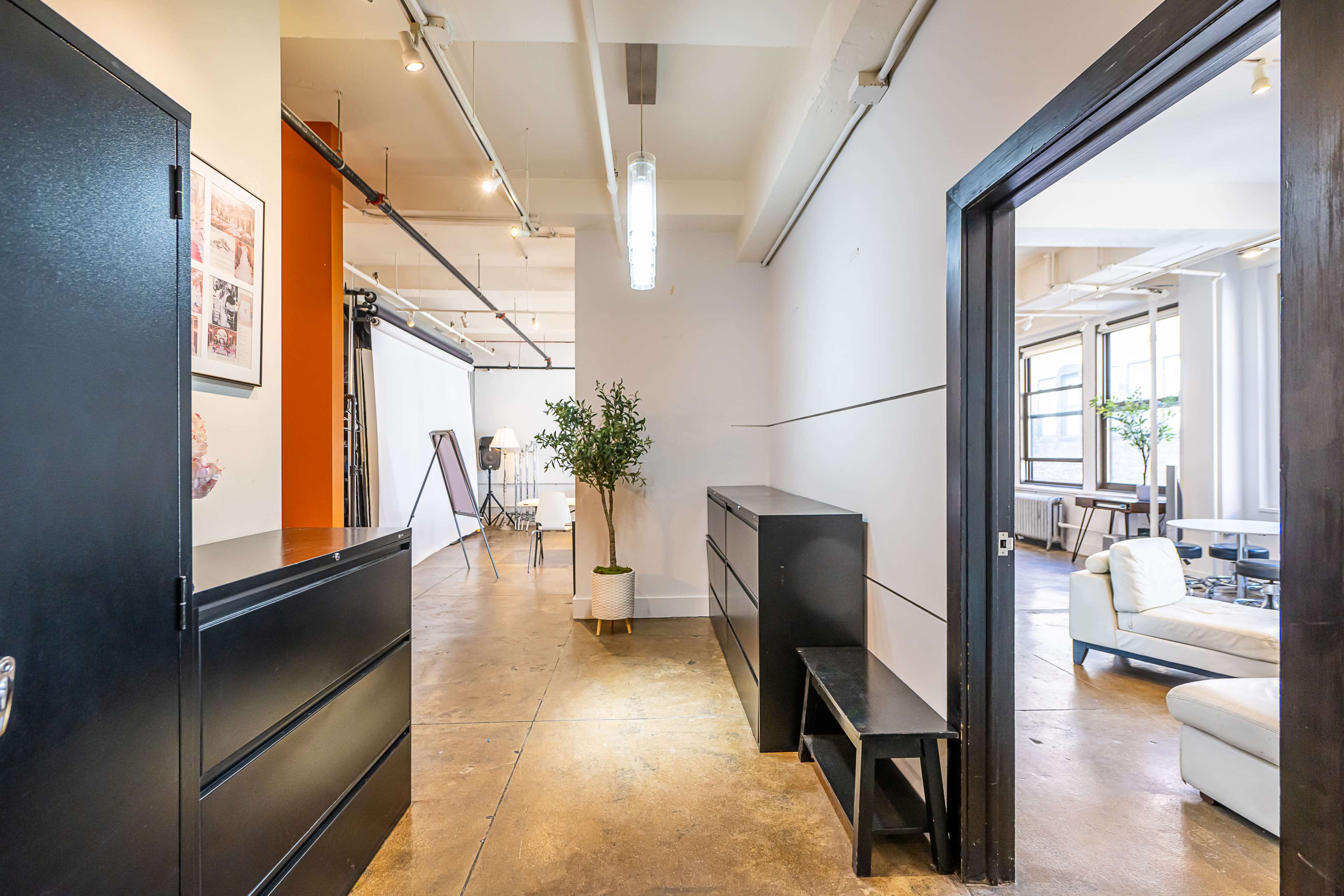 The image shows a hallway leading to a room with black furniture and large windows, featuring a plant and a studio setup in the background.