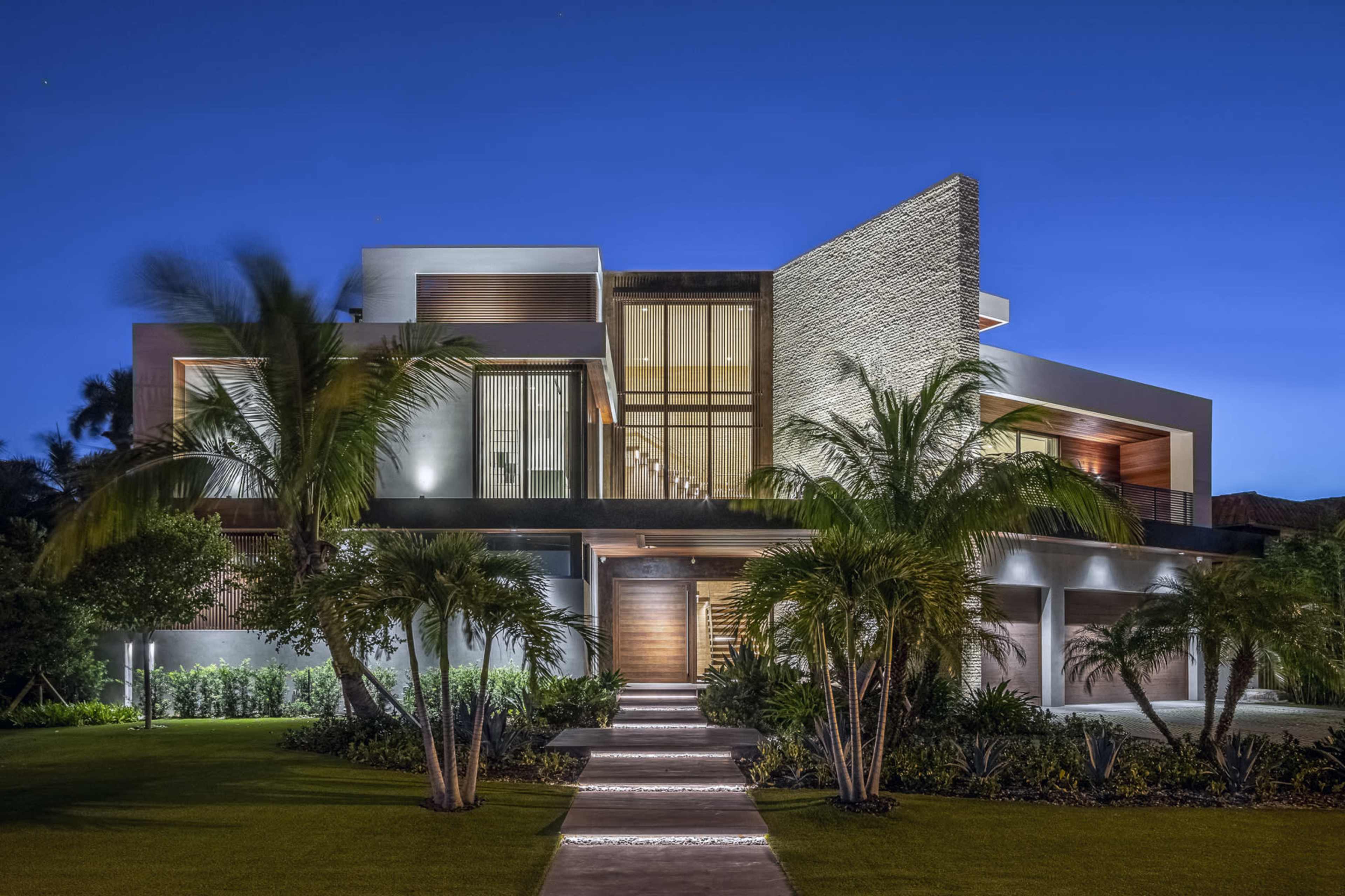 A modern house at night, featuring large windows, a stone facade, and landscaped greenery.