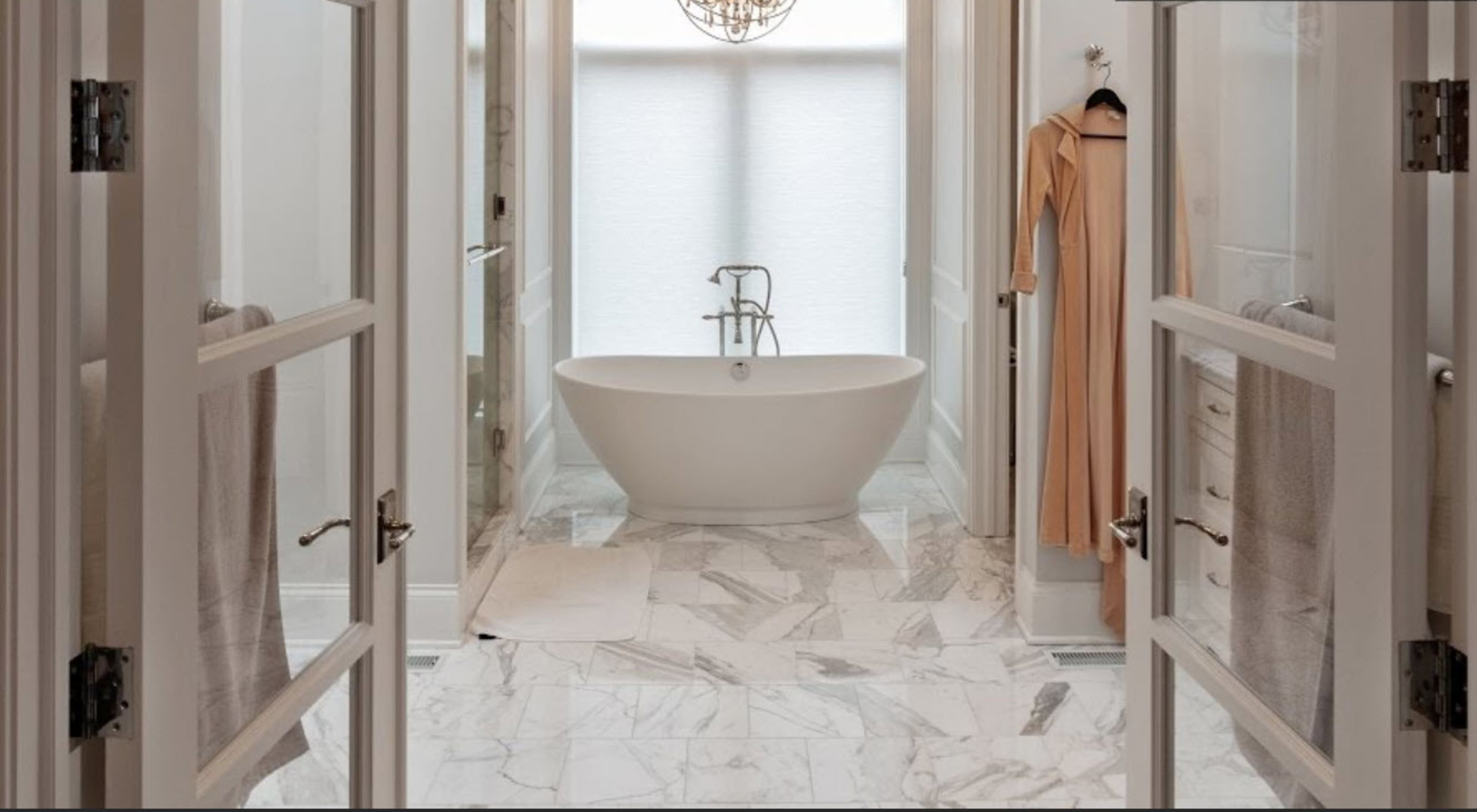 A modern bathroom with a freestanding bathtub centered between two doorways, featuring marble flooring and soft lighting.