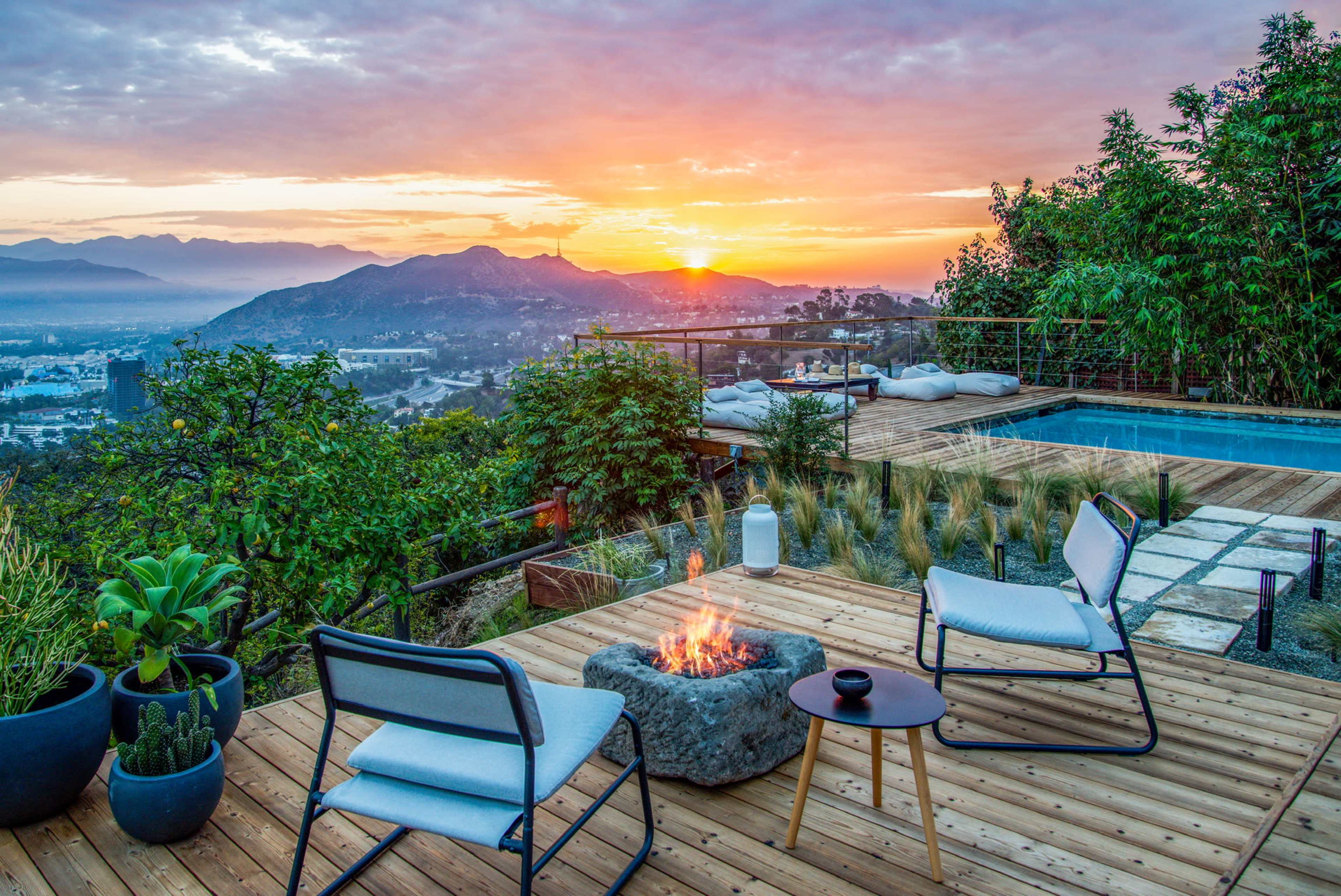 A wooden deck with two chairs and a fire pit overlooks a valley at sunset, framed by lush greenery and a swimming pool.