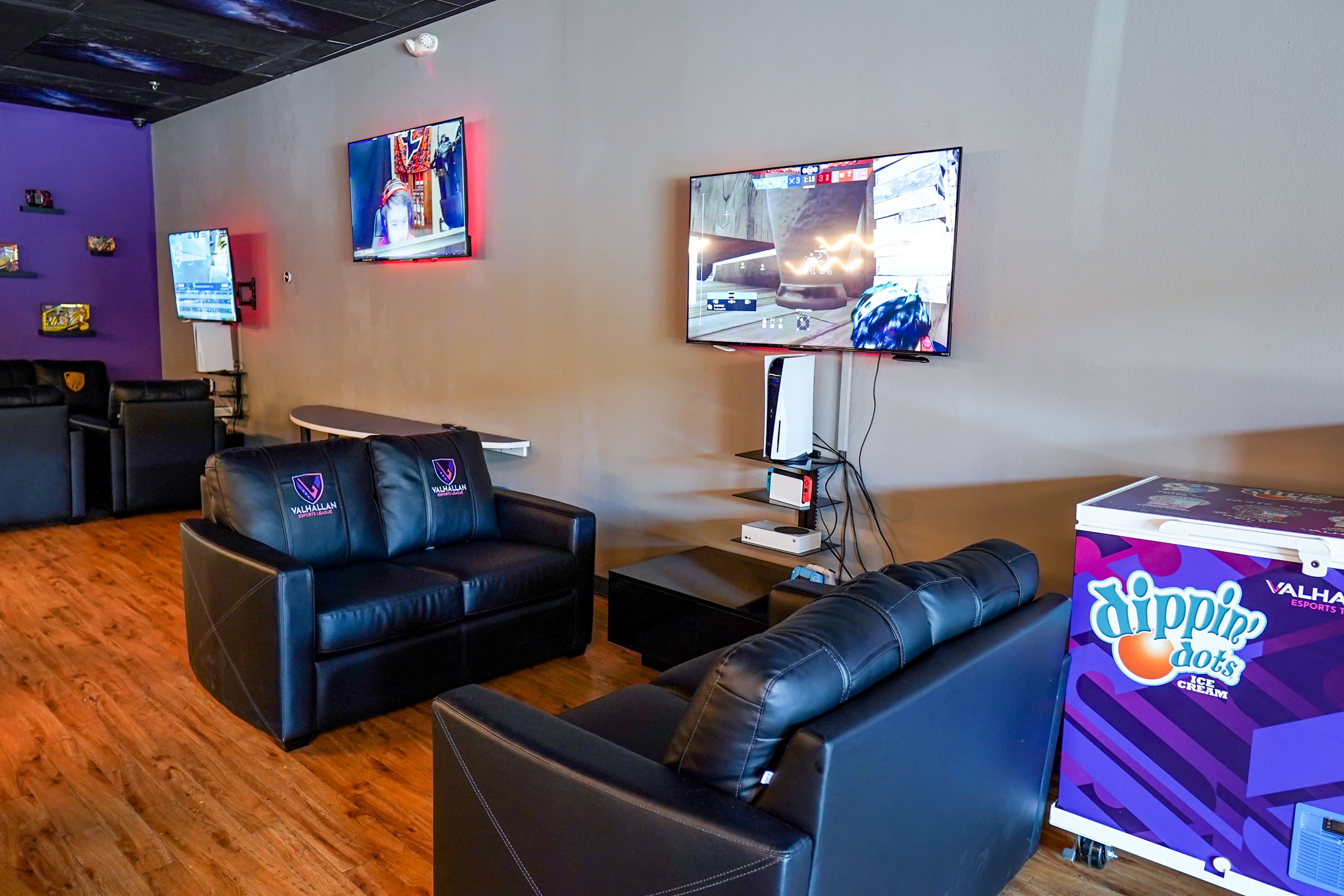 The image shows a modern lounge area with black leather couches, a wall-mounted television displaying a video game, and a colorful ice cream vending machine in the corner.