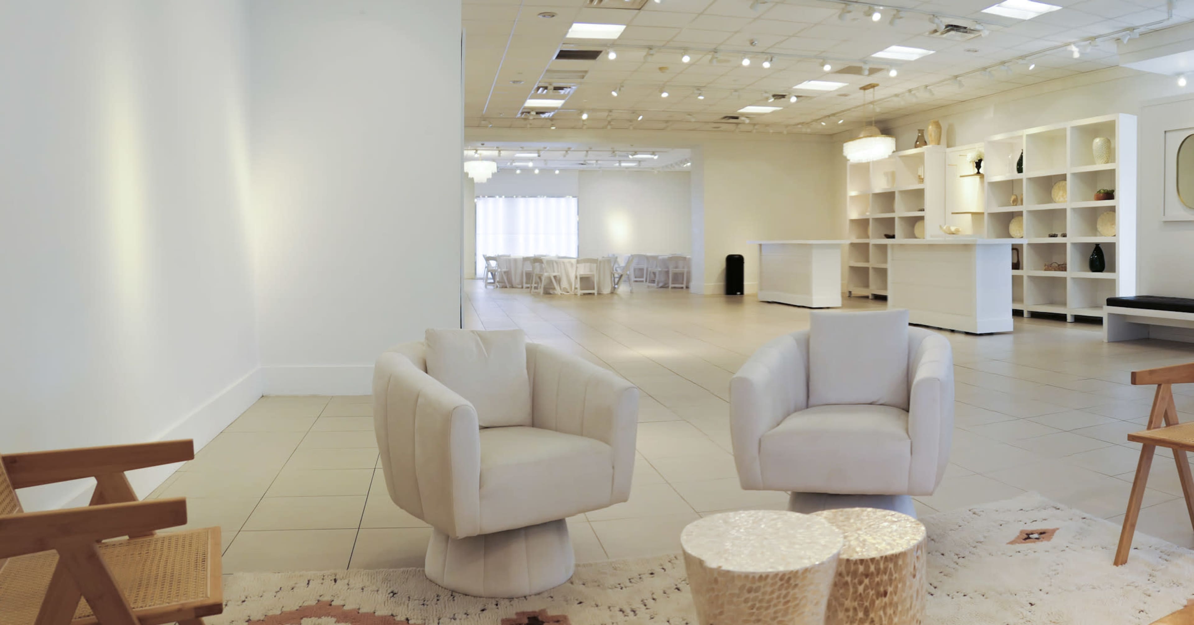 The image shows a spacious, well-lit indoor area featuring two cream-colored armchairs, a small table made of natural materials, and a minimalist design with white walls and shelving.
