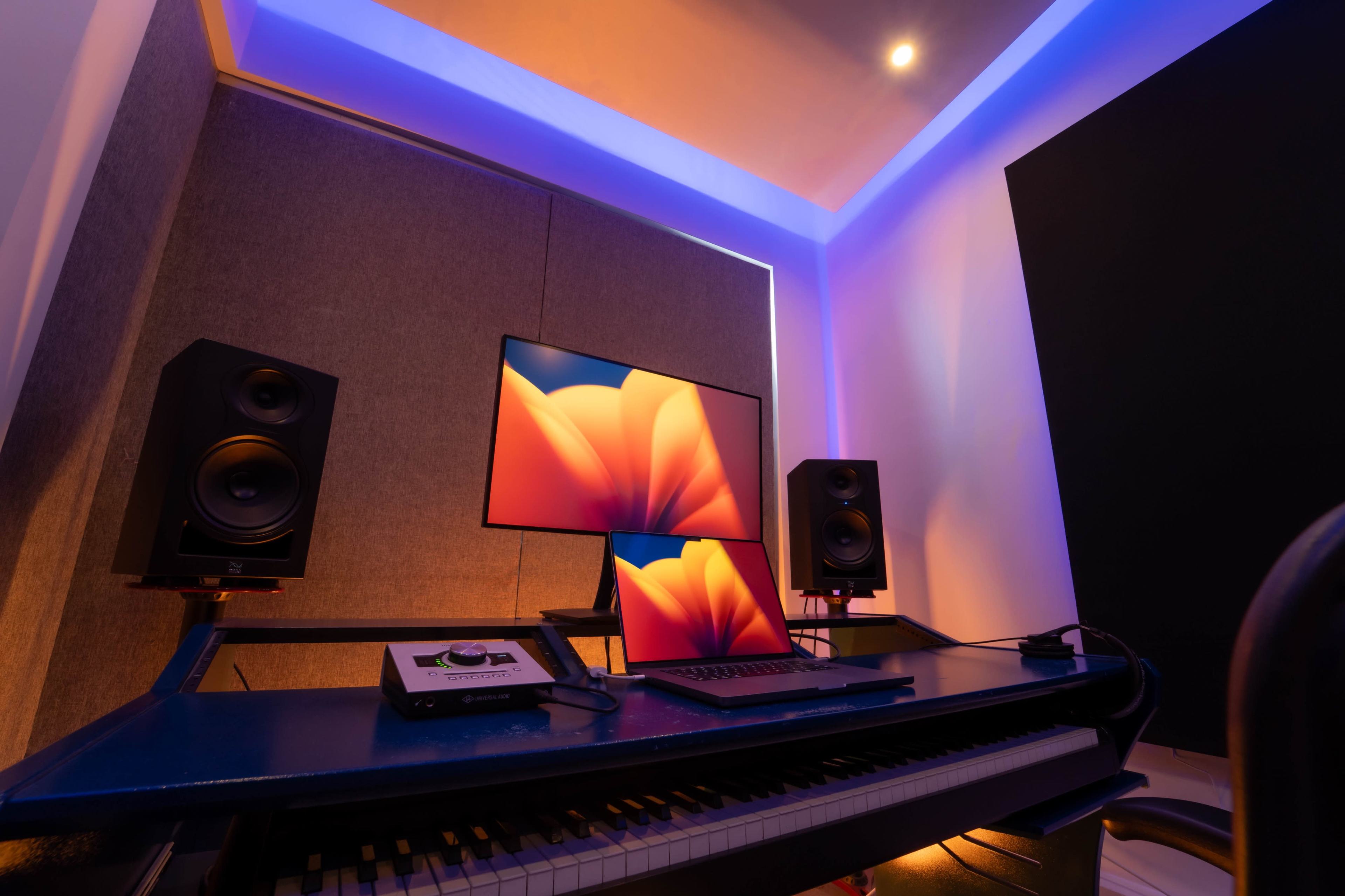 A music production setup featuring a keyboard, two large speakers, a laptop, and a wall-mounted monitor in a dimly lit room with colored lighting.