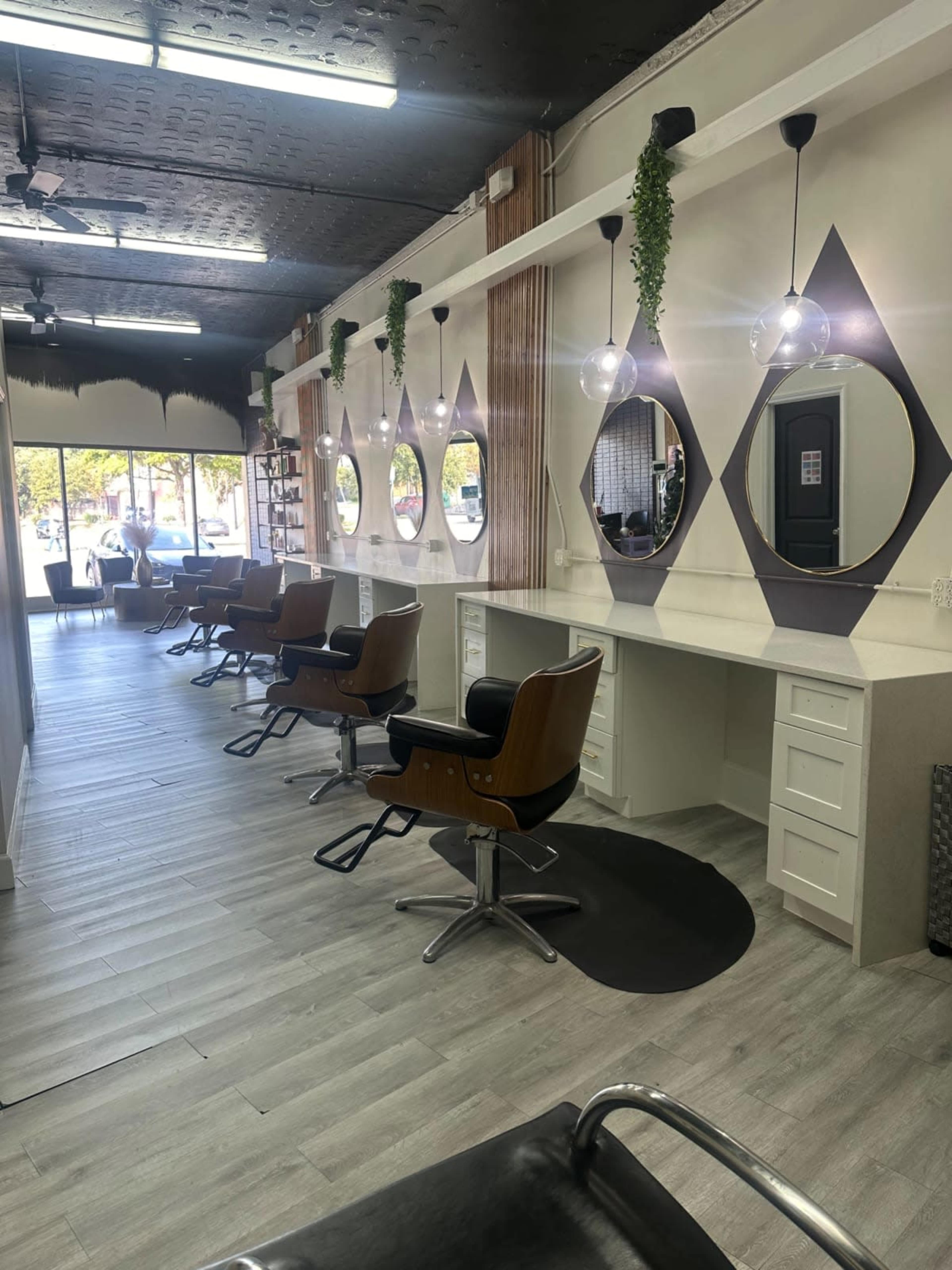The image shows a modern hair salon with several styling stations, each featuring a mirror and a barber chair, lined up against a well-lit wall that leads to a windowed entrance.