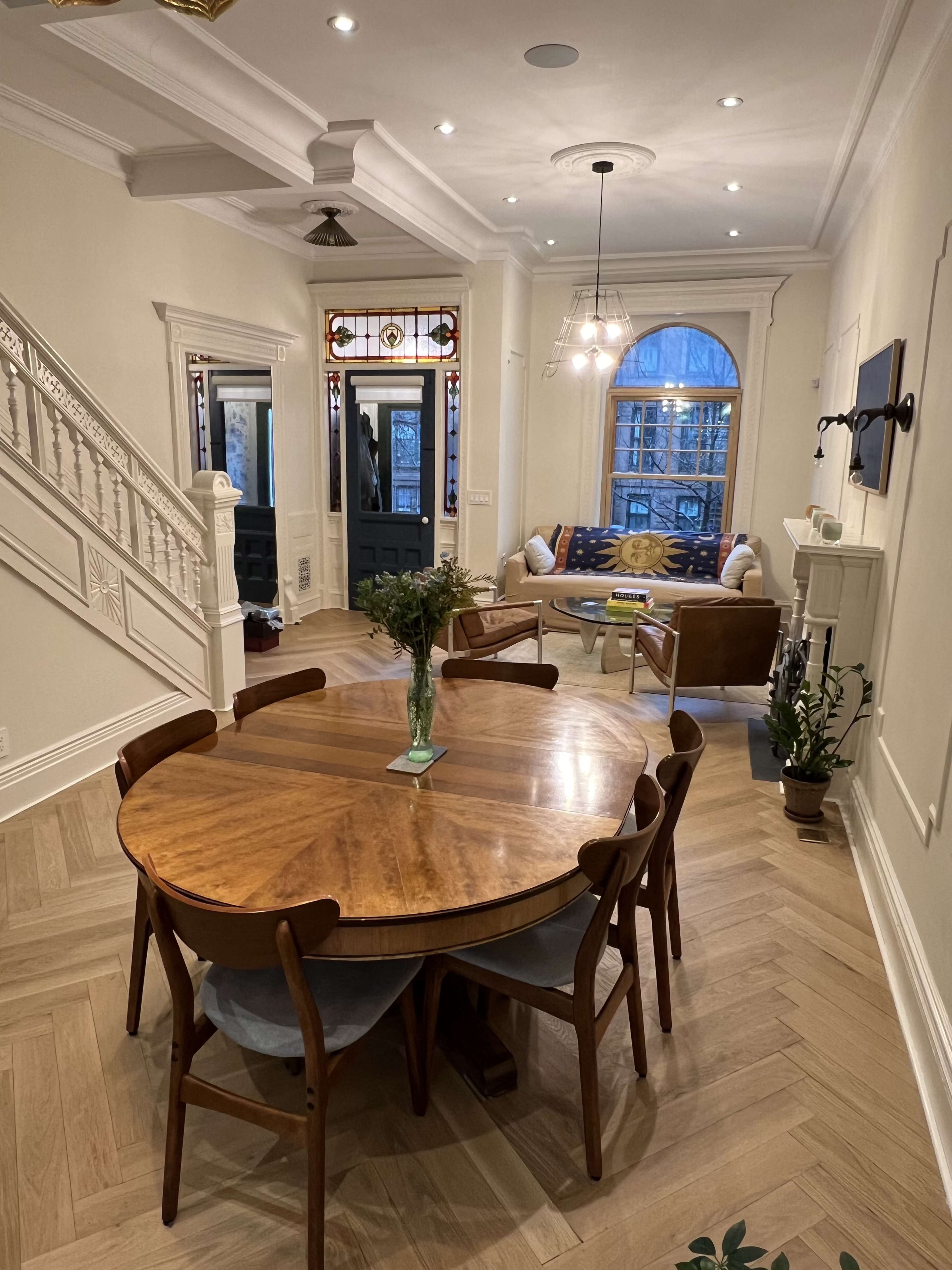 A well-lit, open space featuring a dining area with a wooden table and chairs, leading to a living area with a couch and decorative windows.
