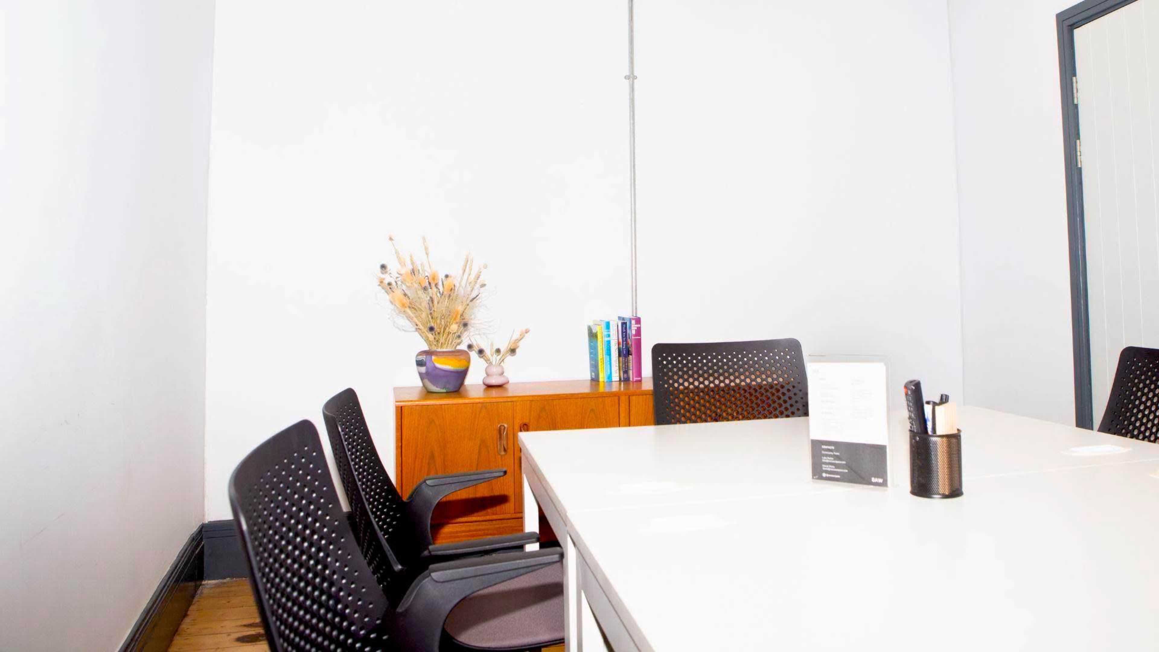 The image shows a conference room with a white wall, a wooden sideboard, a vase of dried flowers, and black chairs around a table.