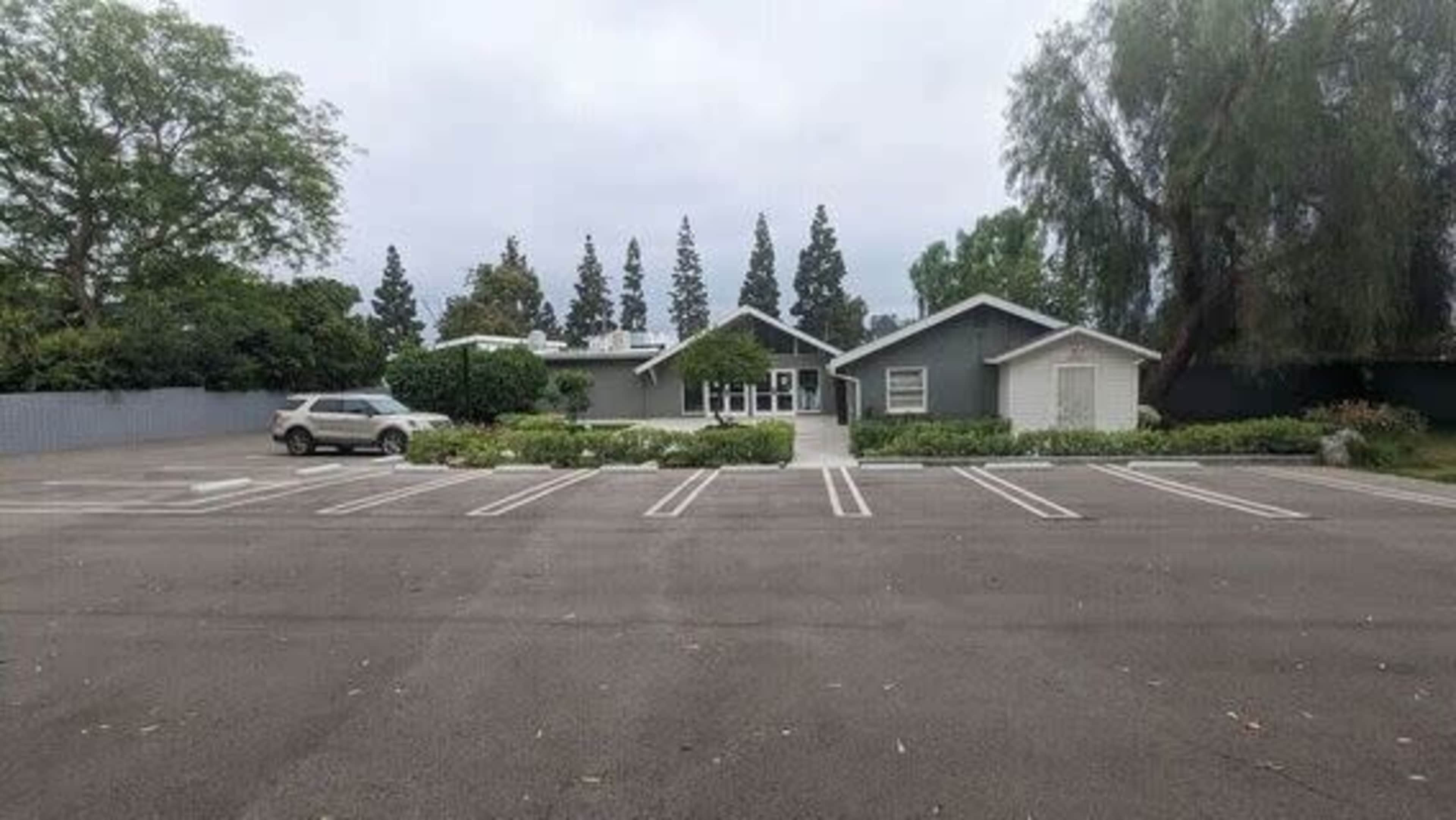 A gray house with a front porch is situated in an empty parking lot lined with marked spaces and surrounded by trees.