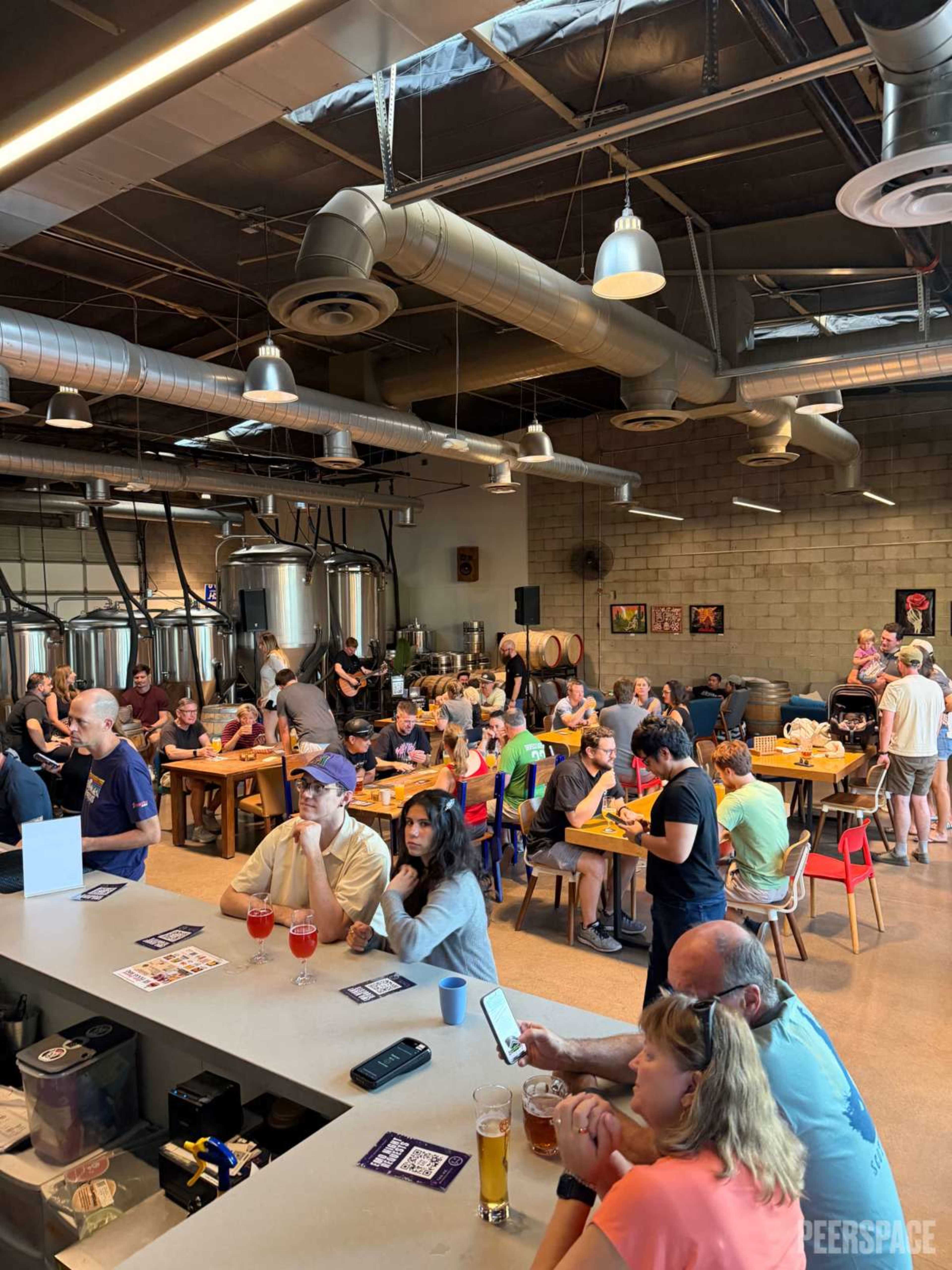 The image shows a lively brewery interior filled with patrons seated at tables, enjoying drinks and food, while a stage area is visible in the background.