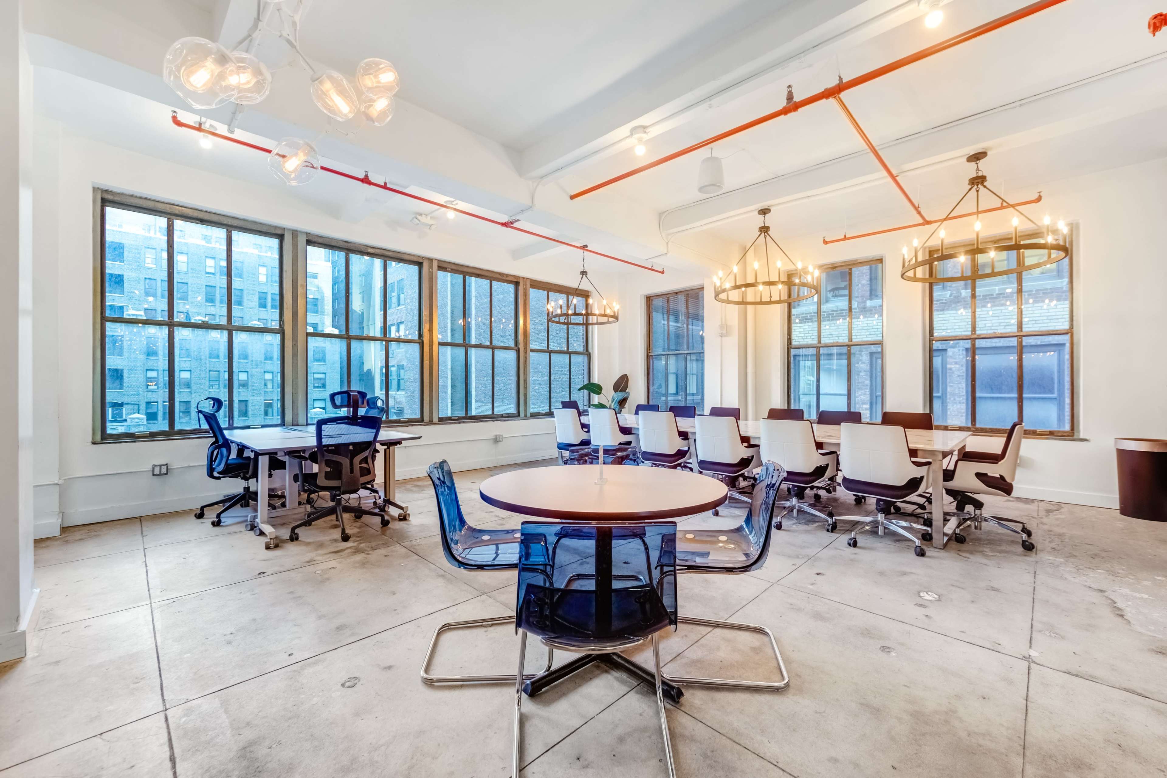 The image shows a modern office space with large windows, several tables, and chairs arranged for meetings, illuminated by overhead light fixtures.