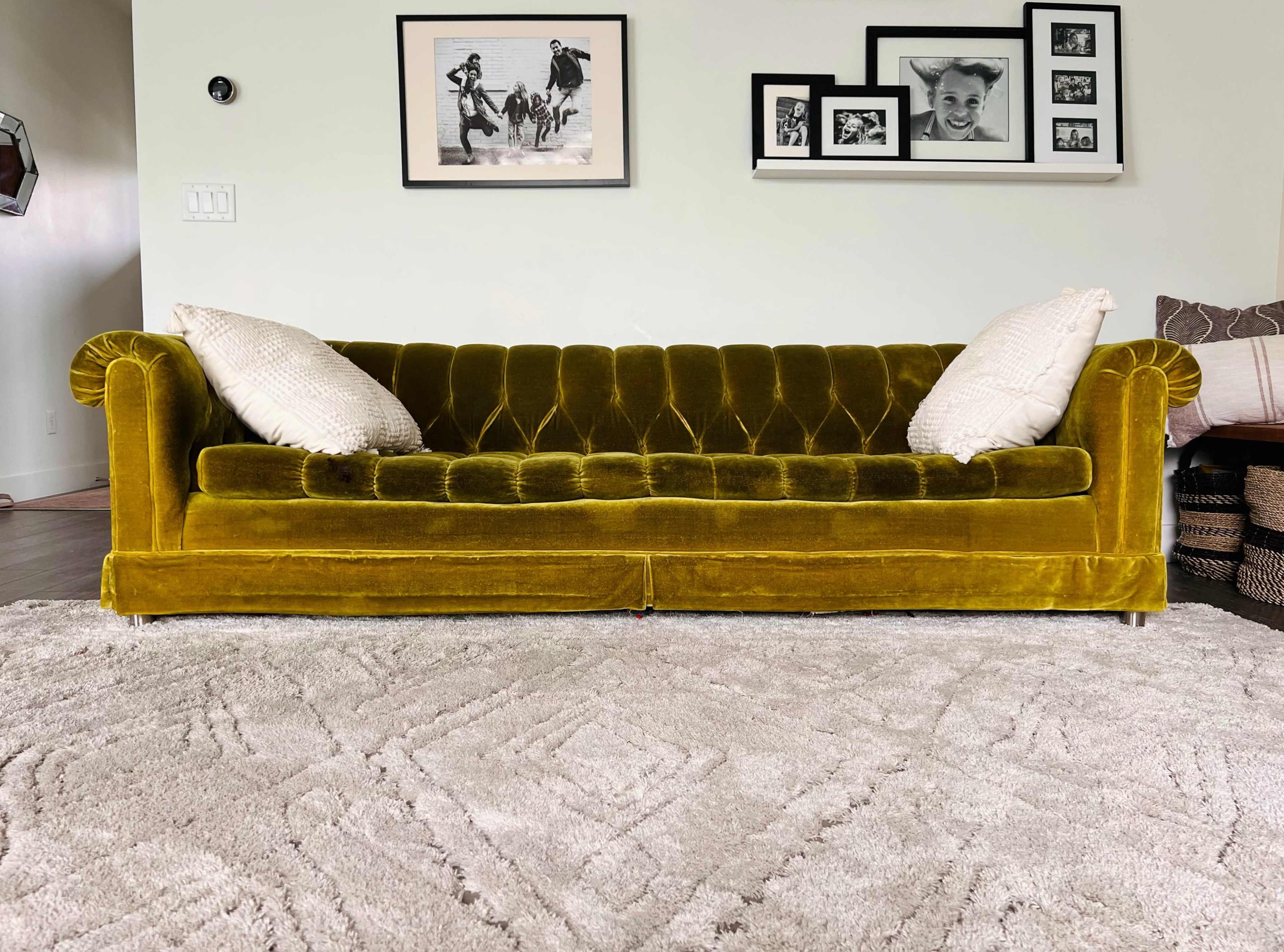 A velvet yellow sofa with tufted upholstery and two white decorative pillows, placed on a textured light-colored rug in a room with framed photographs on the wall.