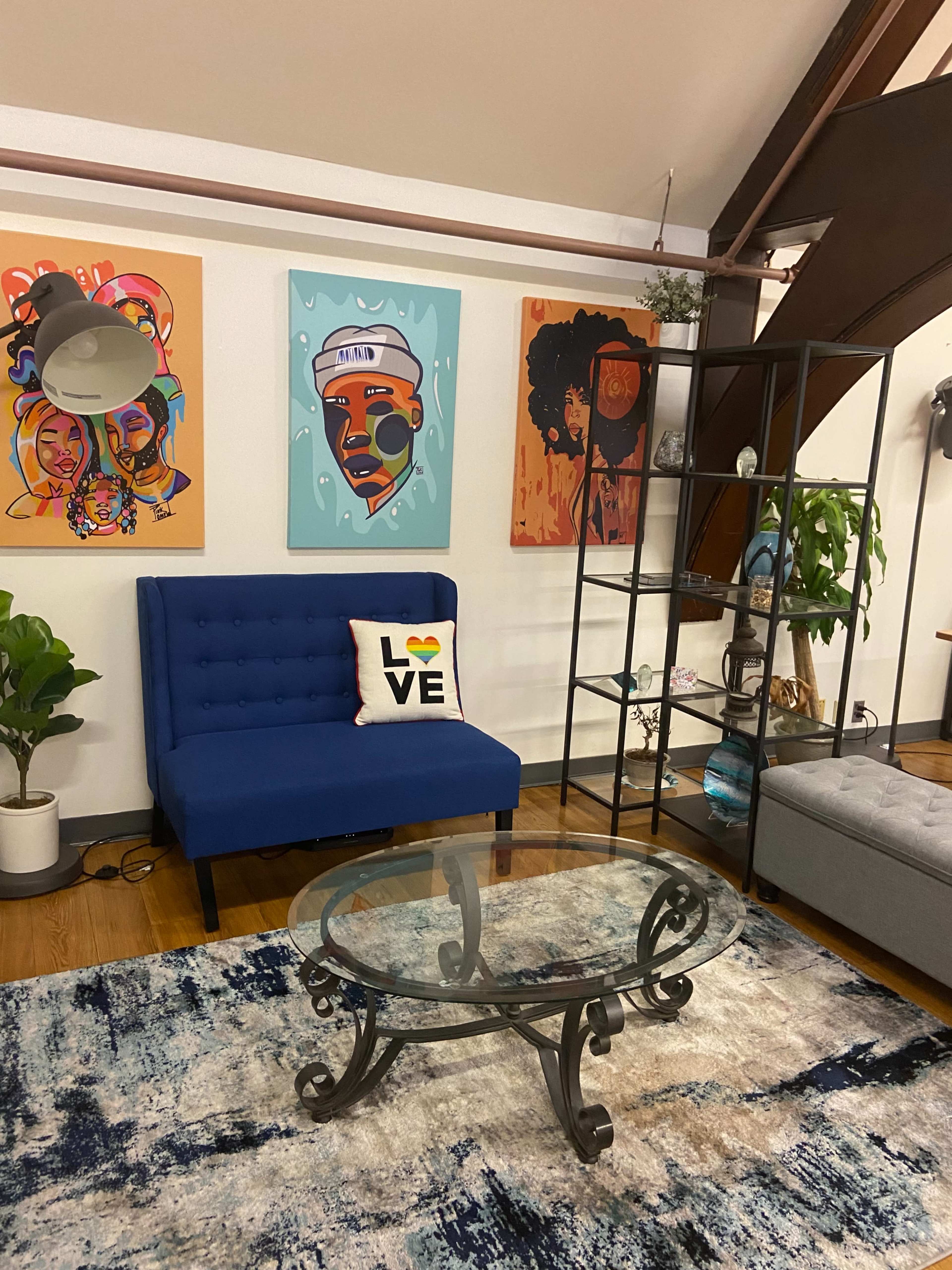 A blue couch with a "LOVE" pillow, a glass coffee table, and colorful artwork displayed on the walls in a modern living room setting.